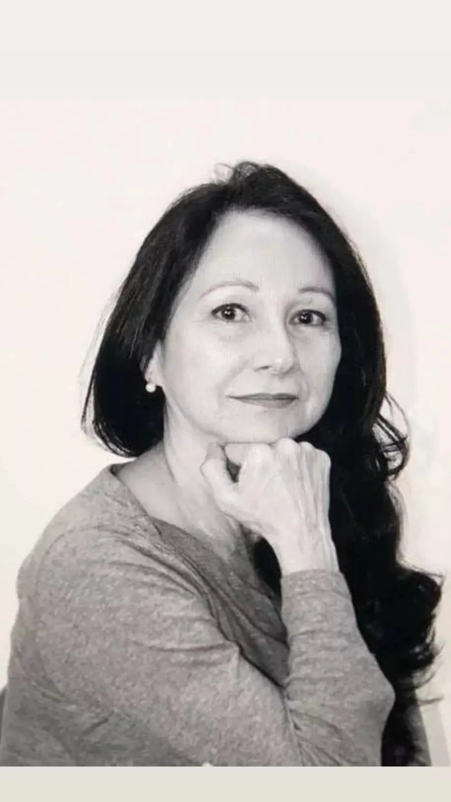 Black and white portrait of a woman with long dark hair, resting her chin on her hand, wearing a long-sleeve top and pearl earrings.