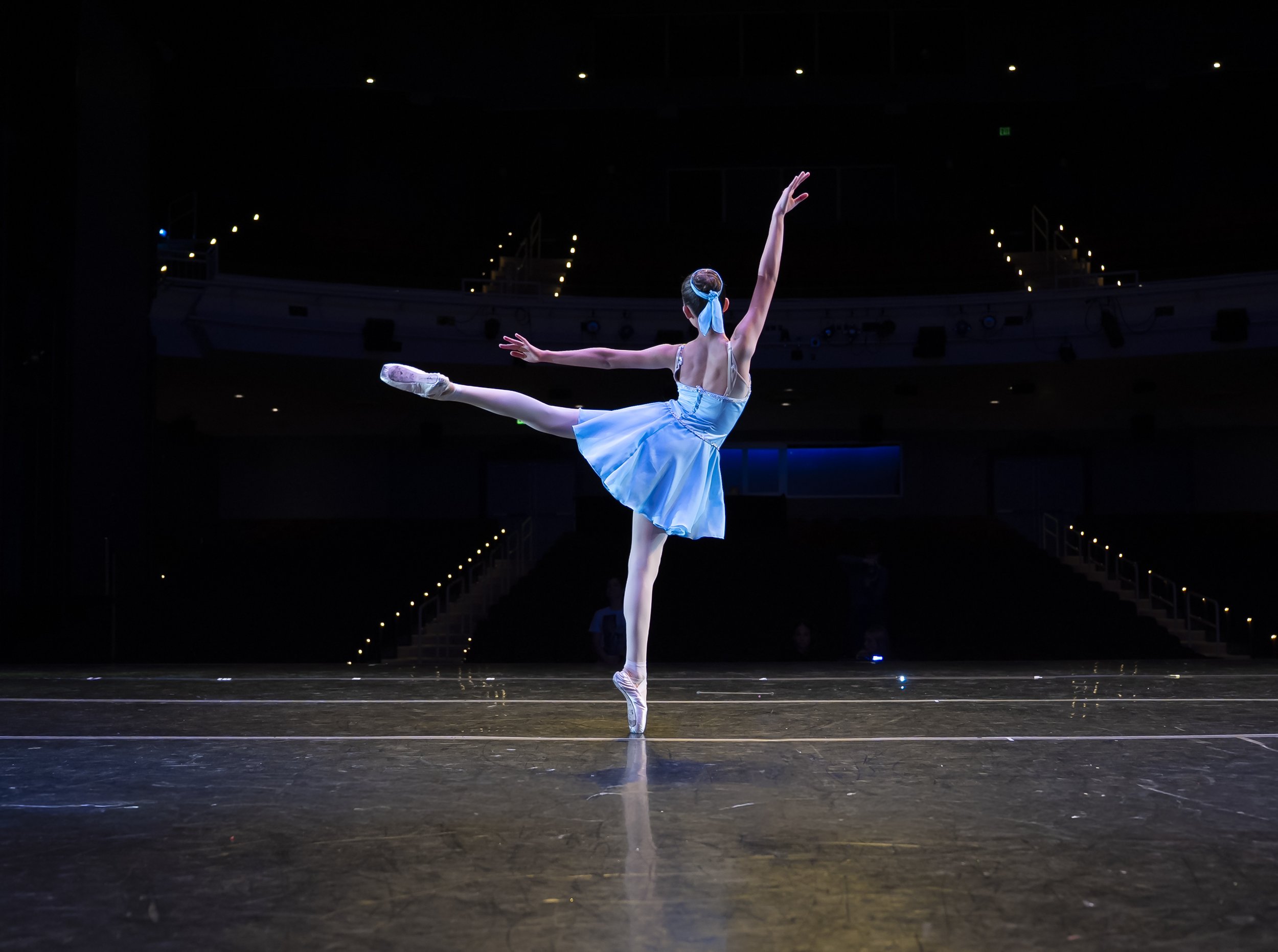 A ballet dancer in a blue tutu and matching headband performing an arabesque on stage with a dark background and stage lights.