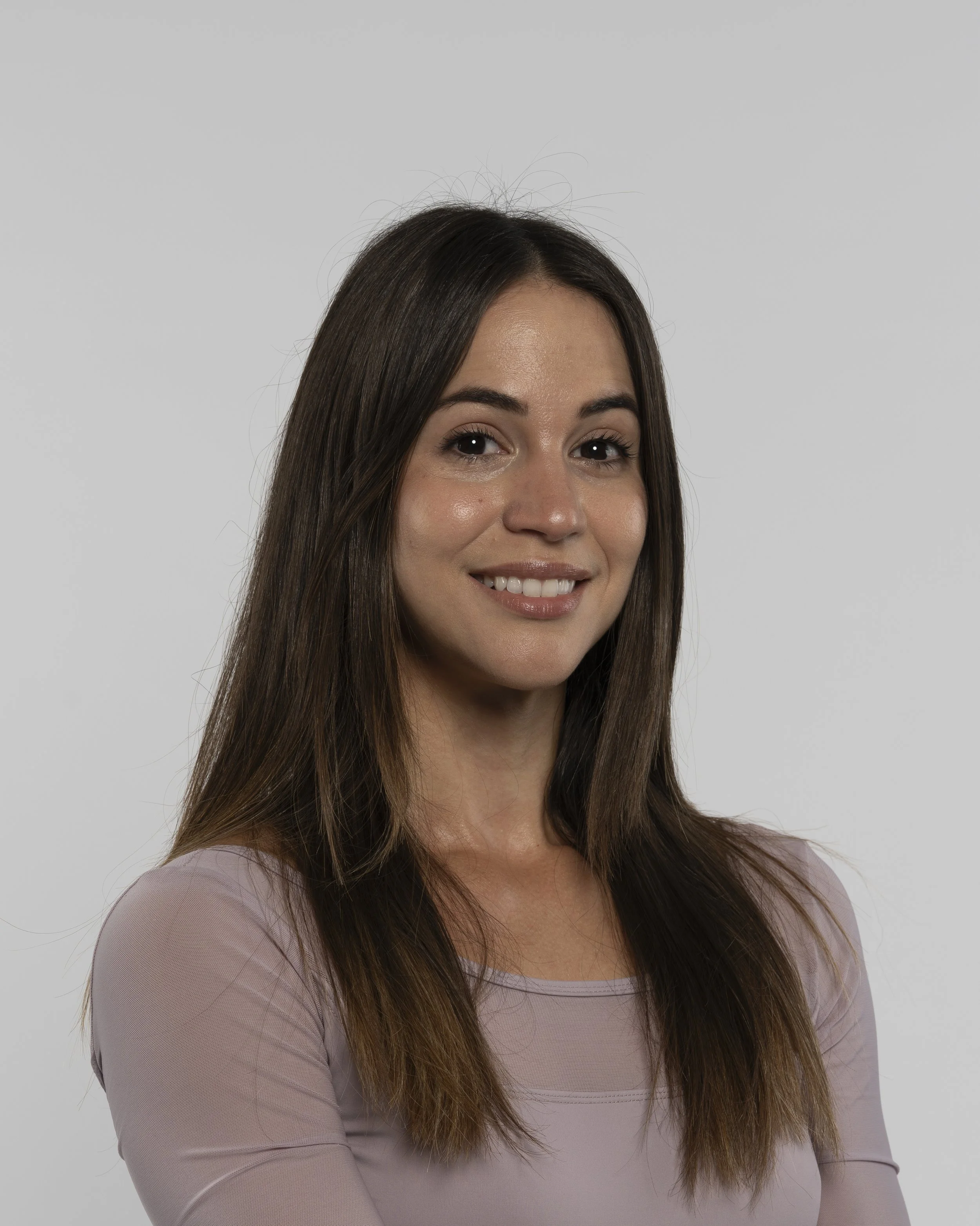 Close-up of a smiling woman with long brown hair, wearing a light pink top, against a plain light background.