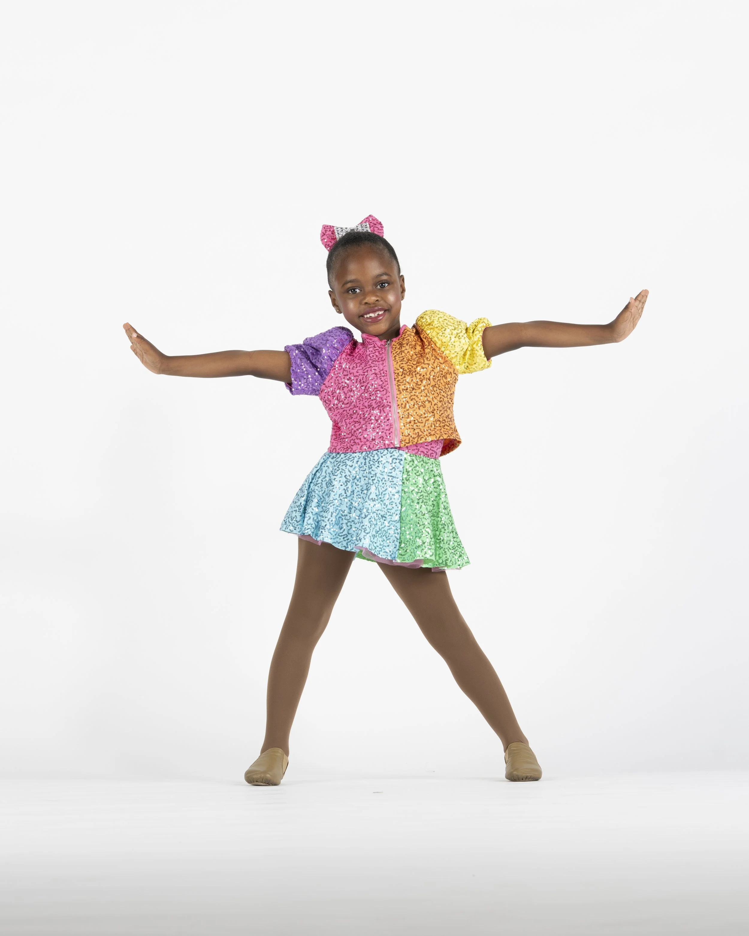 Young girl in a colorful, rainbow-patterned dance costume with arms outstretched, neutral background.