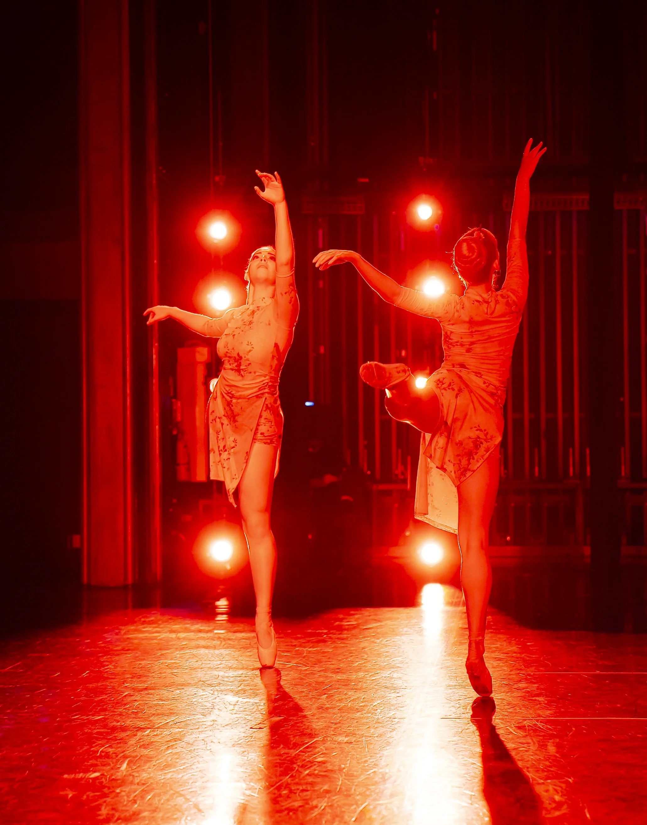 Two ballet dancers performing on stage under intense red lighting, with the background displaying vertical structural elements.