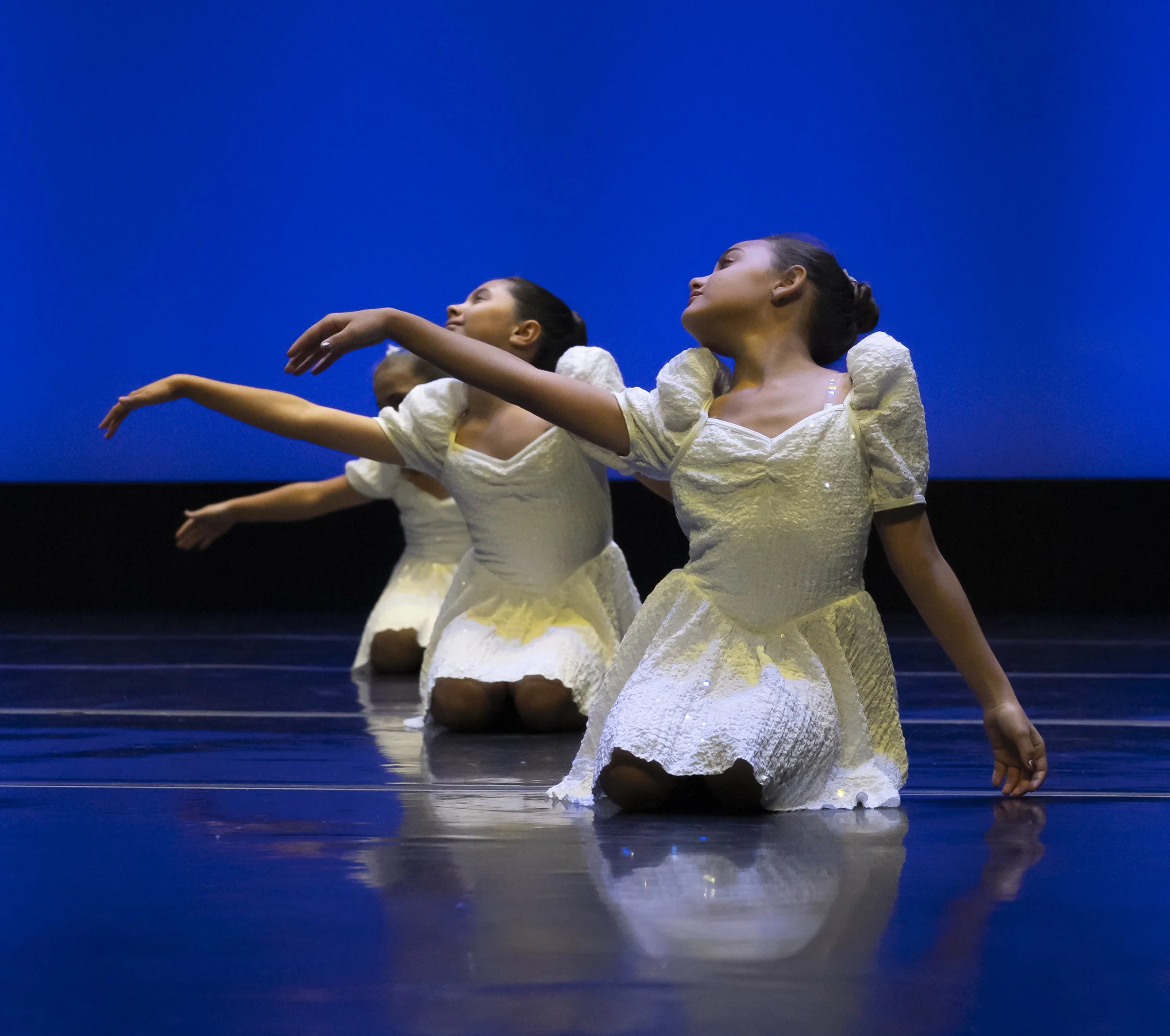 Three girls dressed in white ballet costumes kneeling on a dark stage with their arms extended and heads tilted back, performing a ballet dance against a plain blue background.