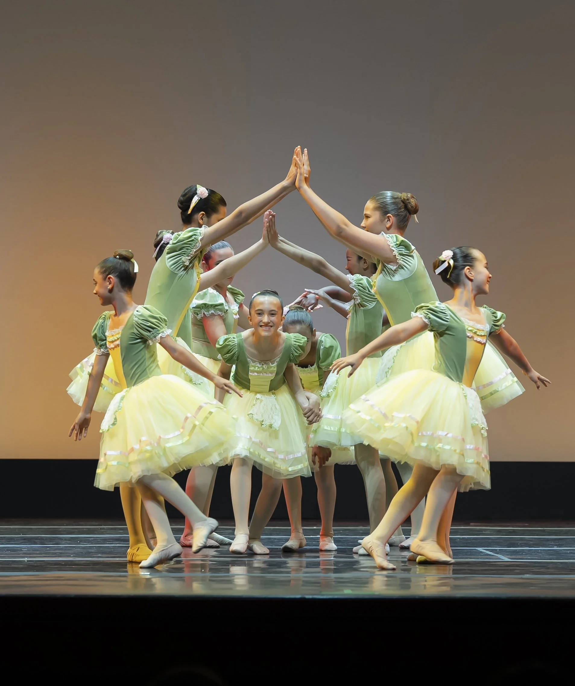 Group of young ballet dancers in pastel green costumes practicing on stage with some raising their arms and others smiling.