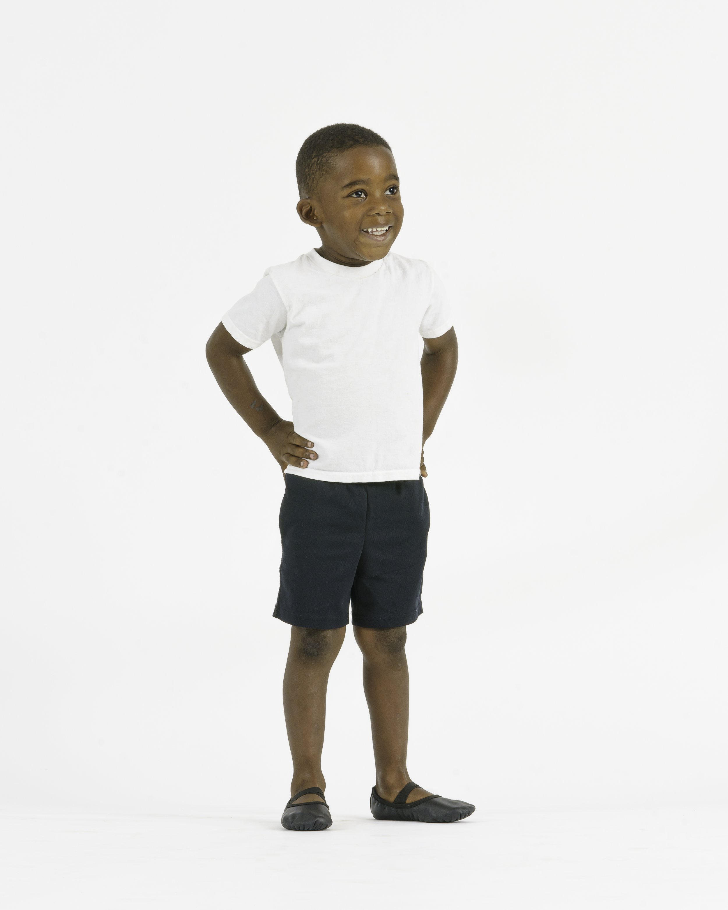 Young boy standing with hands on hips, smiling, wearing a white t-shirt, dark shorts, and black slippers, against a plain white background.