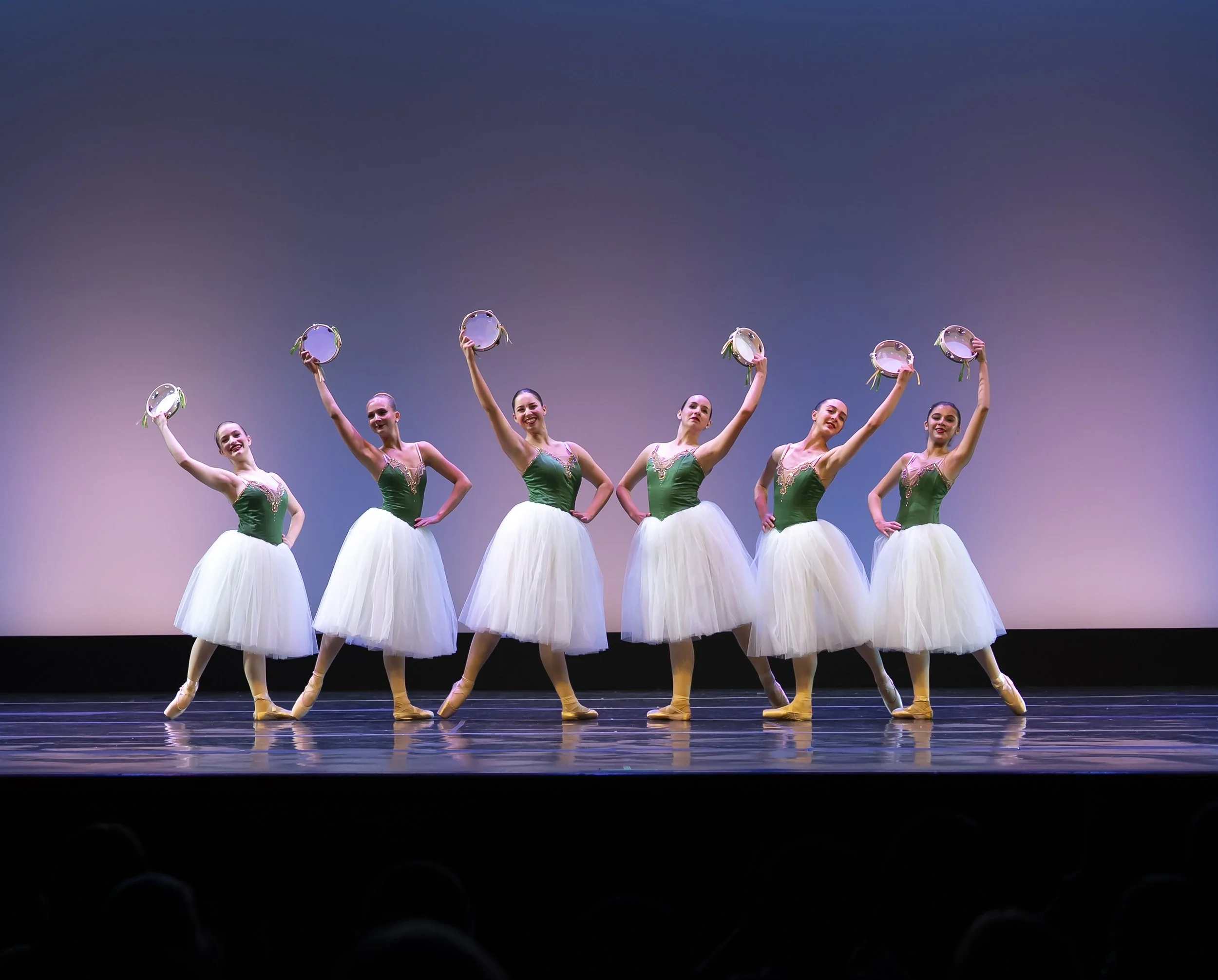 A group of six ballet dancers in green costumes holding tambourines above their heads, standing in a line on stage with a gradient background.