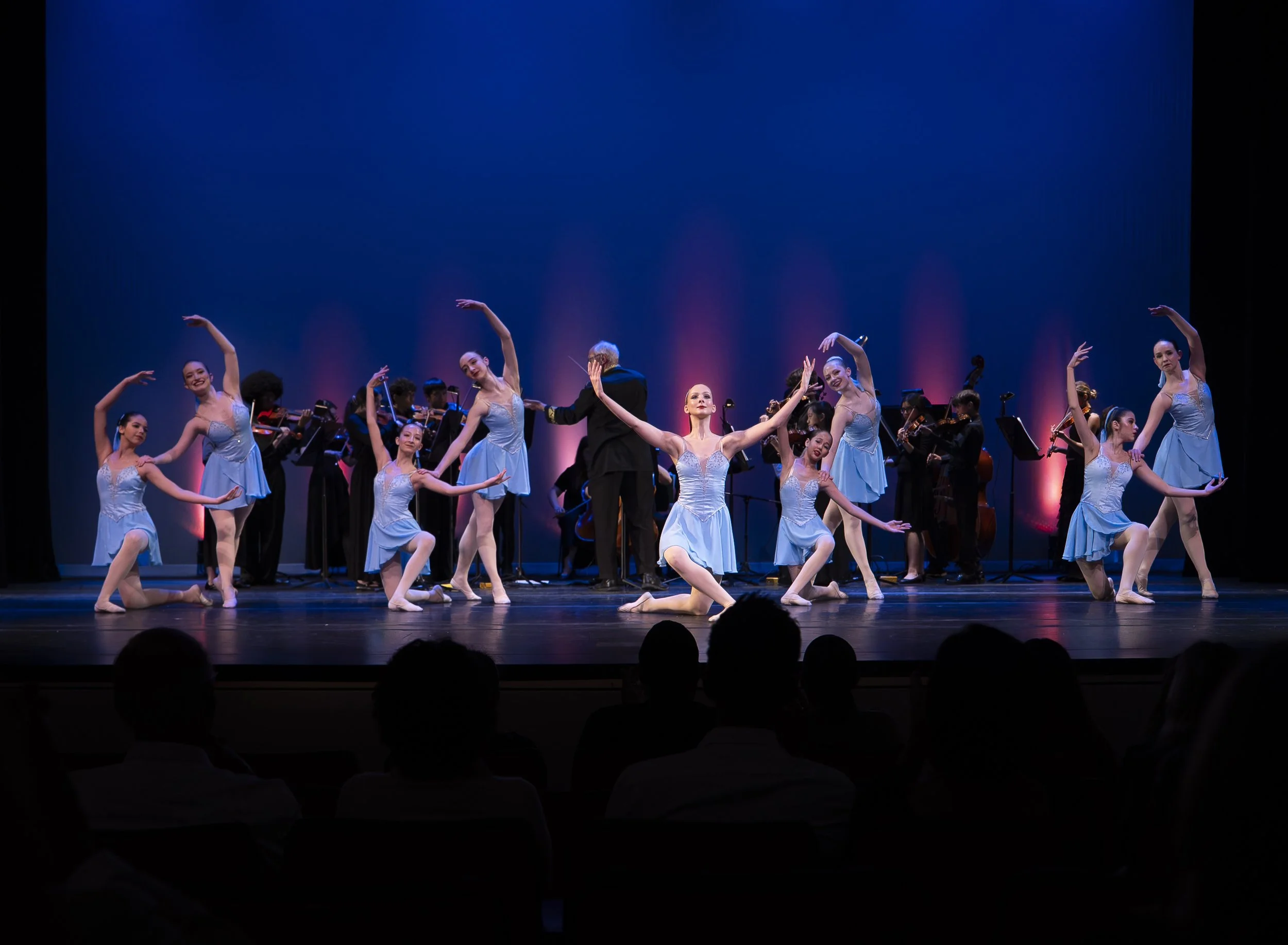 Ballet dancers performing on stage with string musicians in the background, illuminated by pink and blue stage lighting.