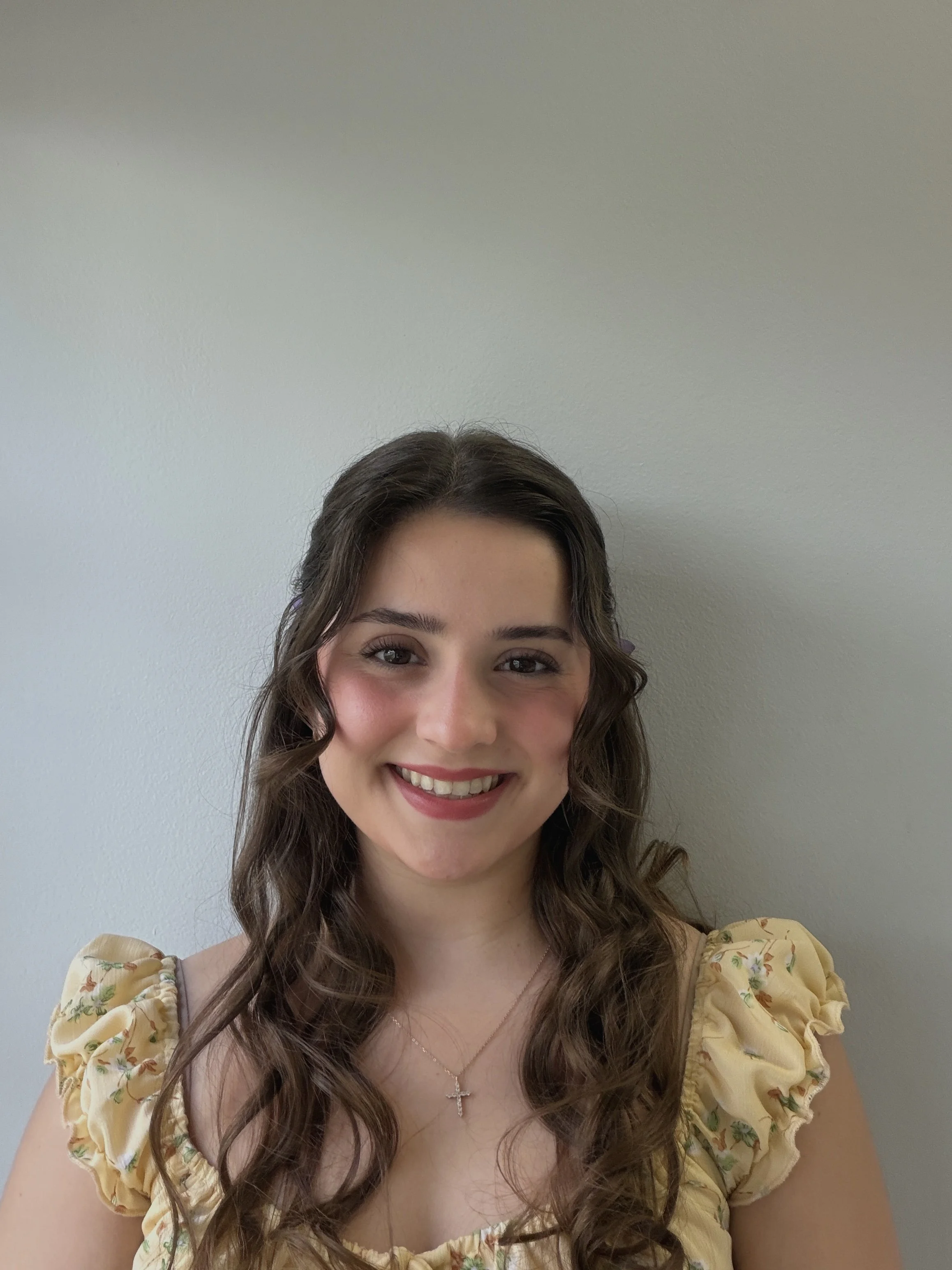 A young woman with long, curly brown hair smiling, wearing a yellow floral blouse and a silver cross necklace, standing against a plain, light gray wall.