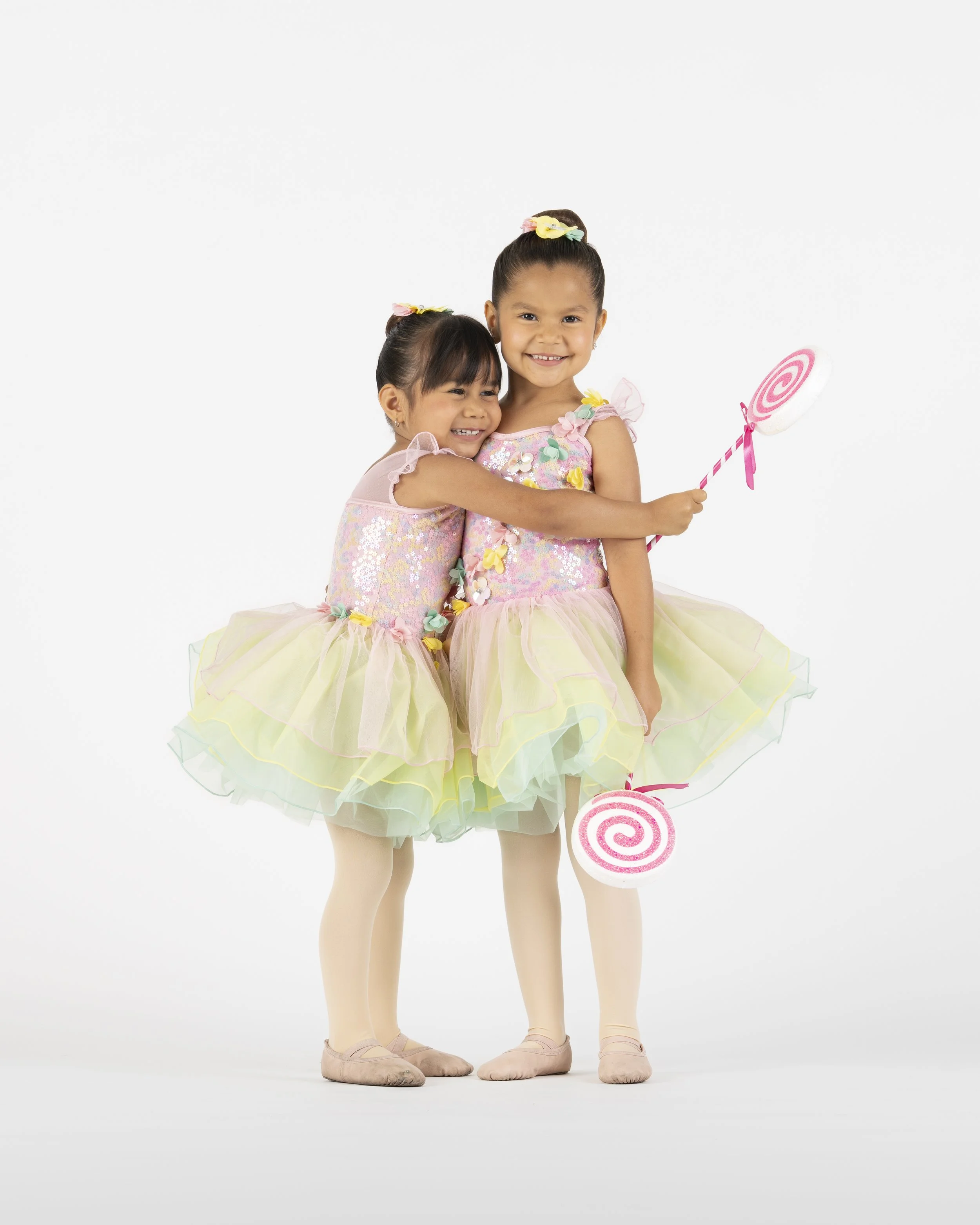 Two young girls dressed as ballerinas in pastel-colored tutus, with one girl holding a pink and white swirled lollipop and smiling at the camera against a plain white background.