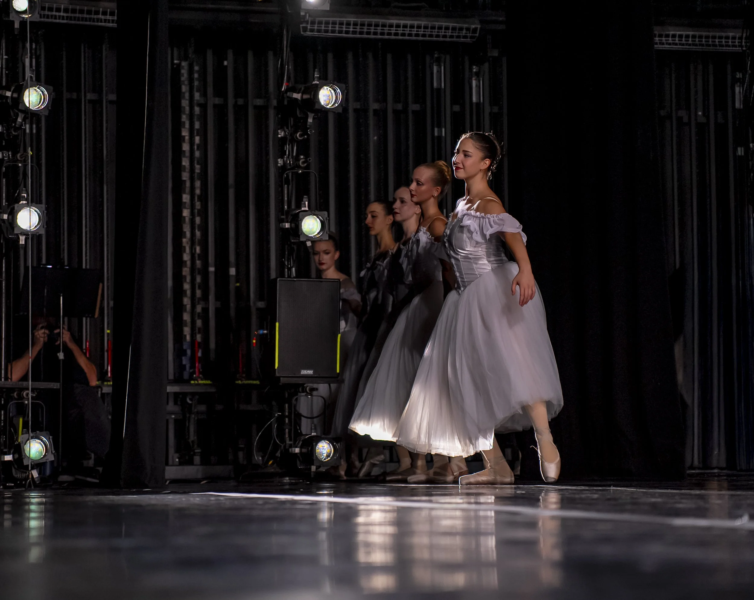 Ballet dancers in ballet costumes practicing on stage, with stage lights and rigging visible in the background.
