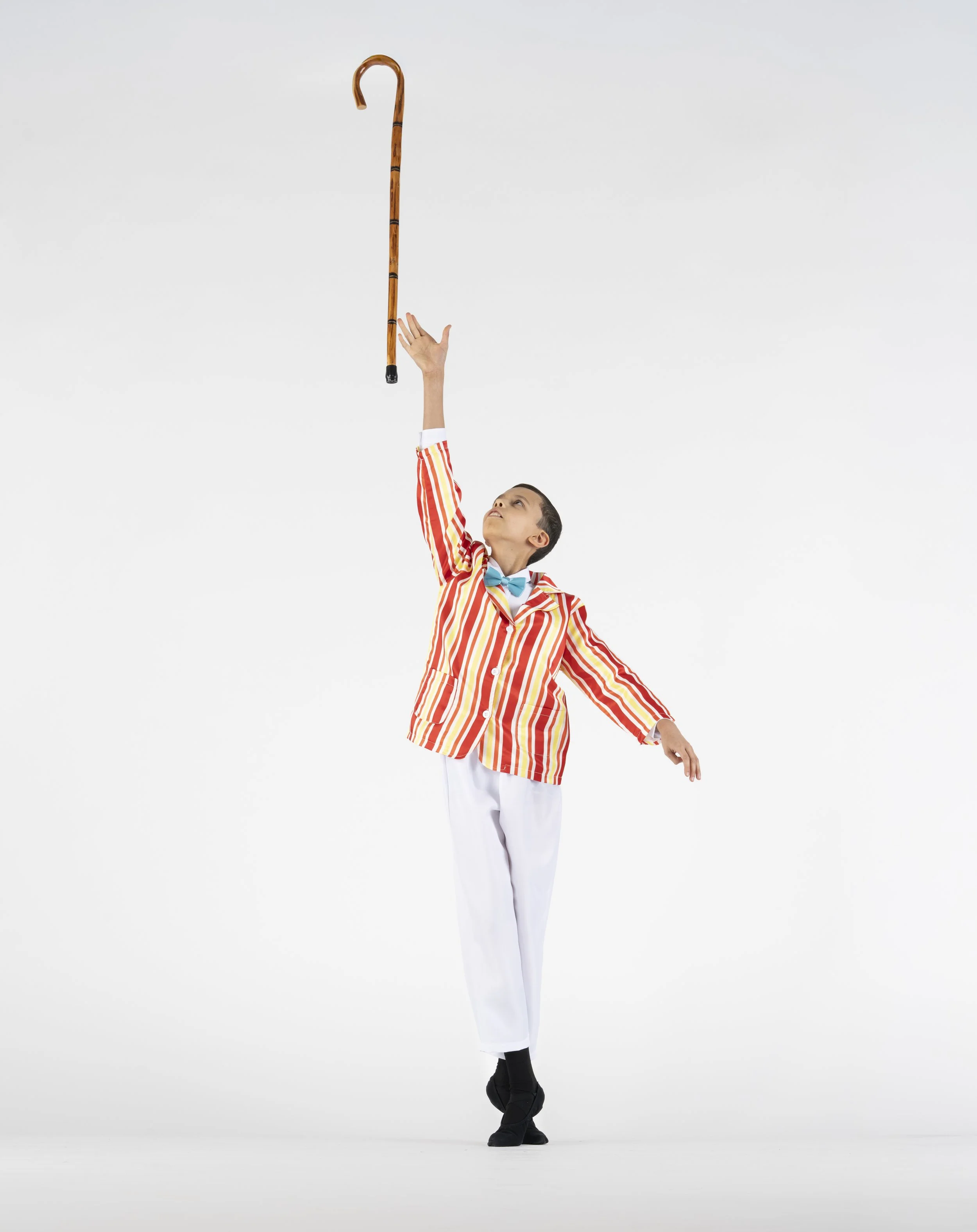 Child in colorful striped blazer and white pants balancing on one leg, holding a wooden cane upright against a plain white background.