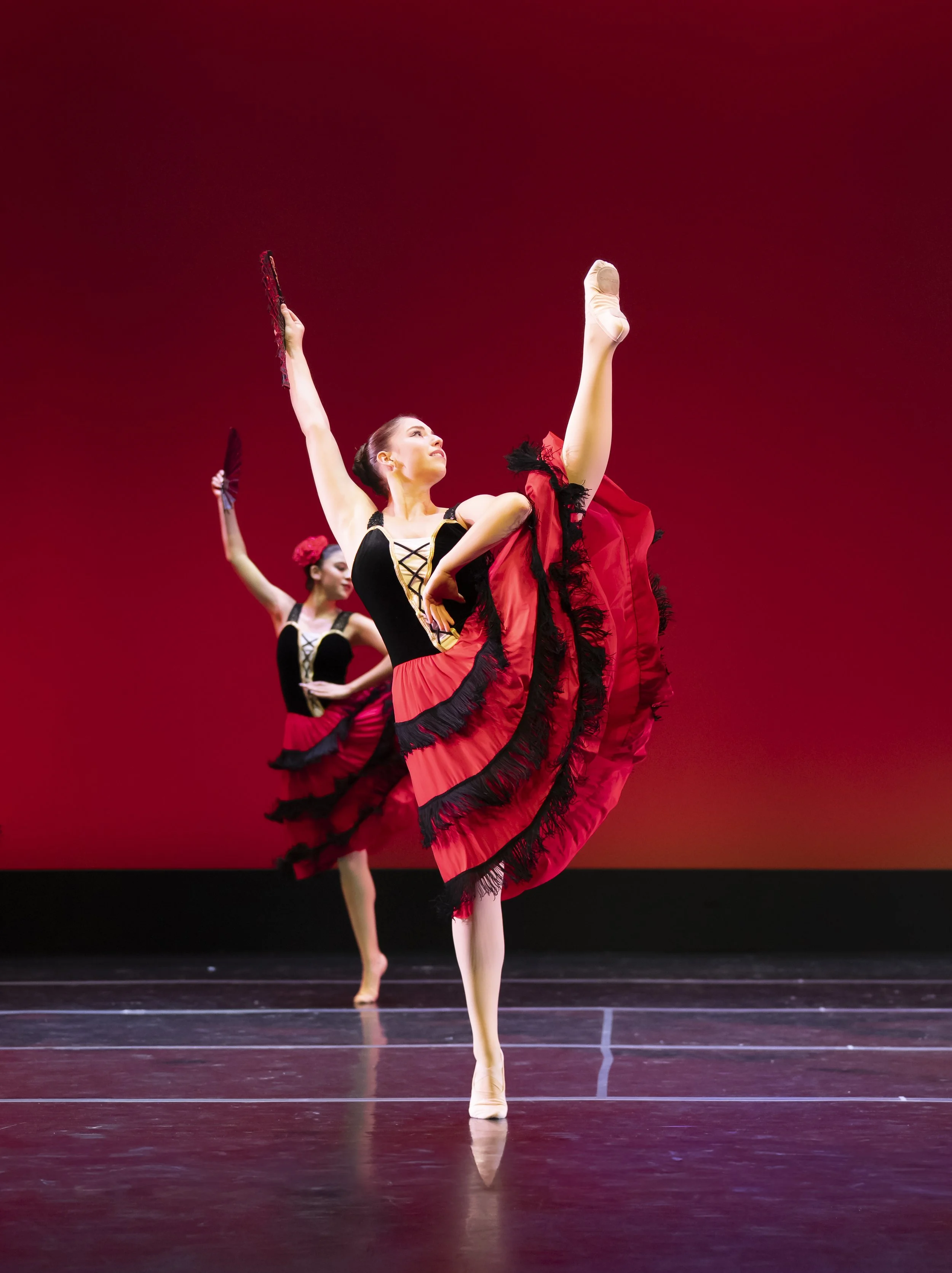 Ballet dancers performing on stage with red background, wearing black and red costumes with ruffles, in ballet poses with one leg raised high.