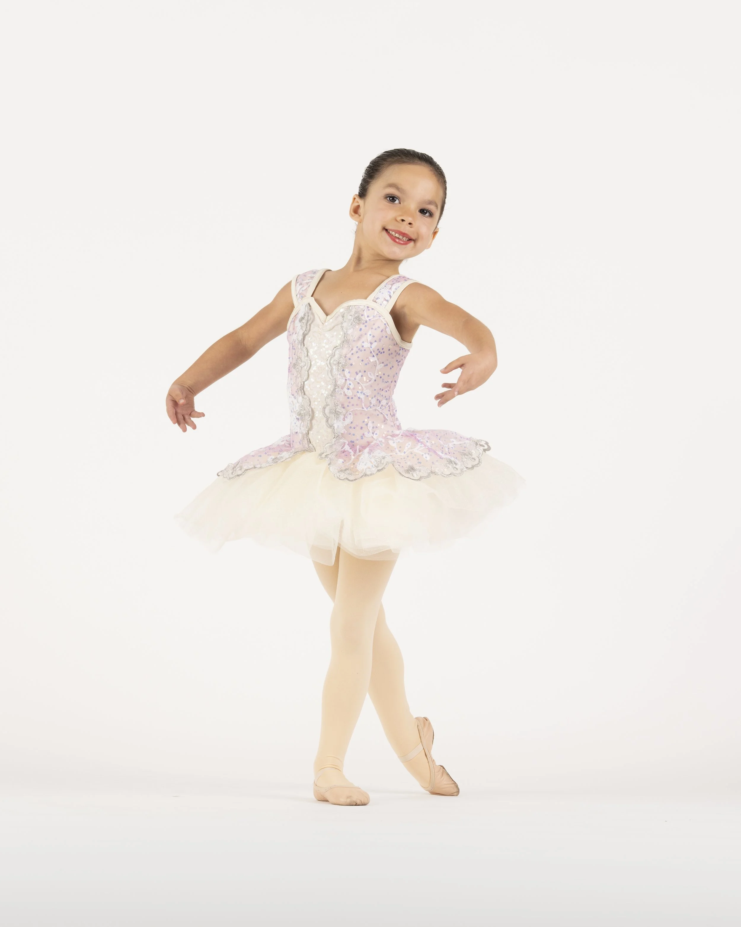 Young girl in a ballet pose wearing a pink and cream tutu, pink ballet slippers, and beige tights, smiling against a white background.