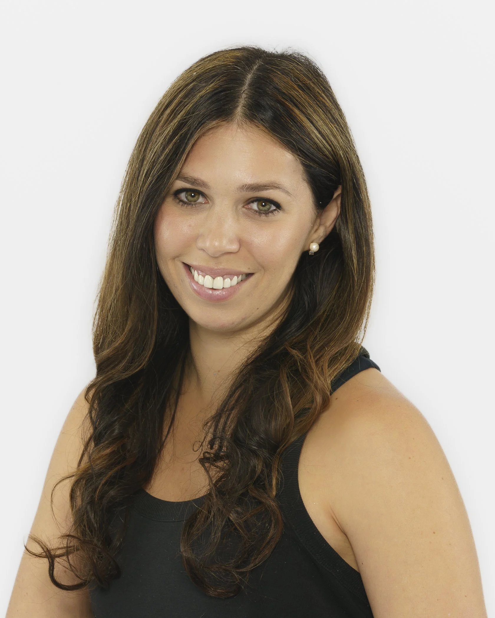 Close-up of a smiling woman with long, wavy brown hair with highlights, wearing pearl earrings and a black sleeveless top, against a white background.