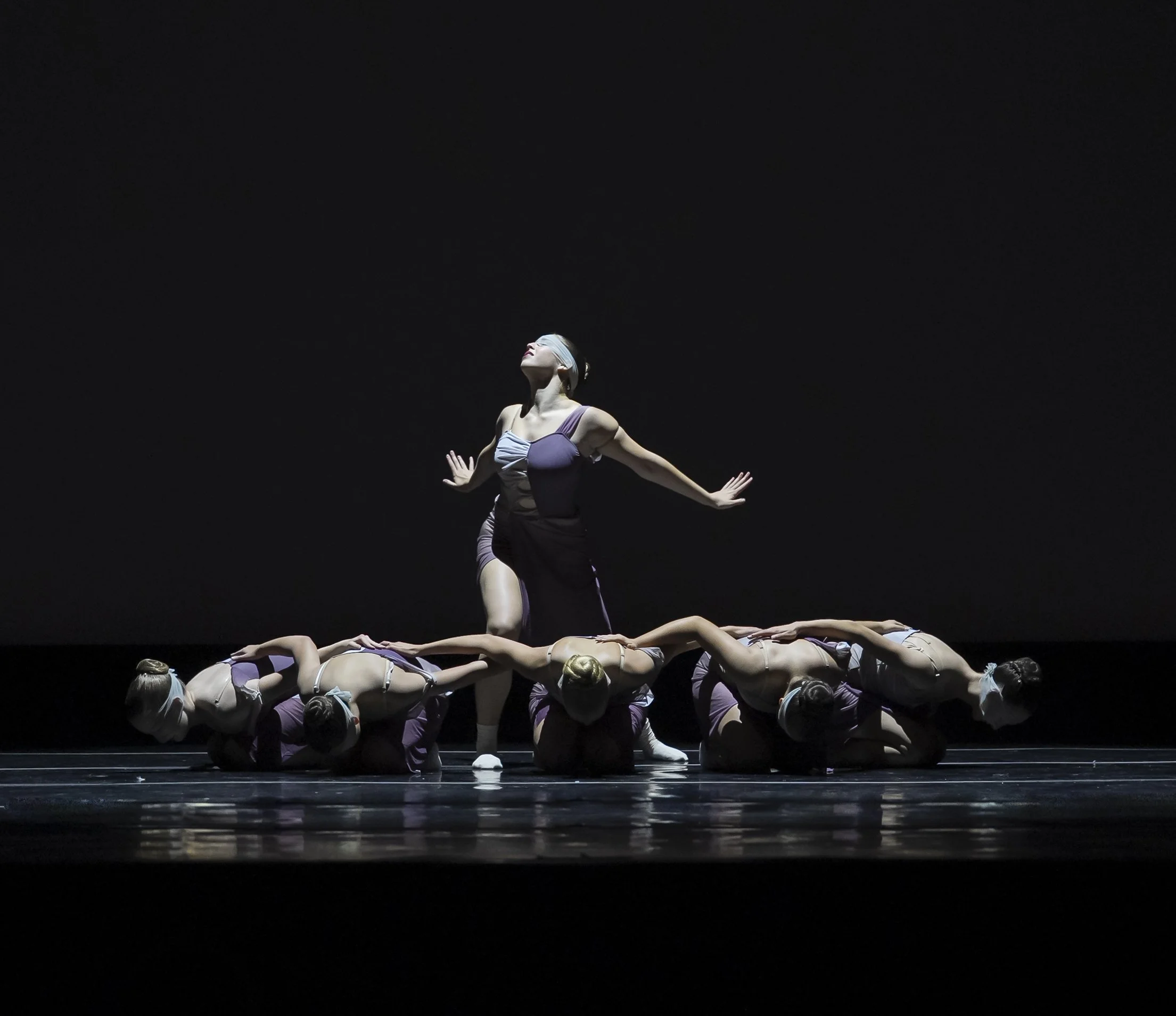 A group of ballet dancers performing a choreographed pose on stage, with one dancer standing upright and others lying on the floor, all wearing purple and beige costumes, against a dark background.