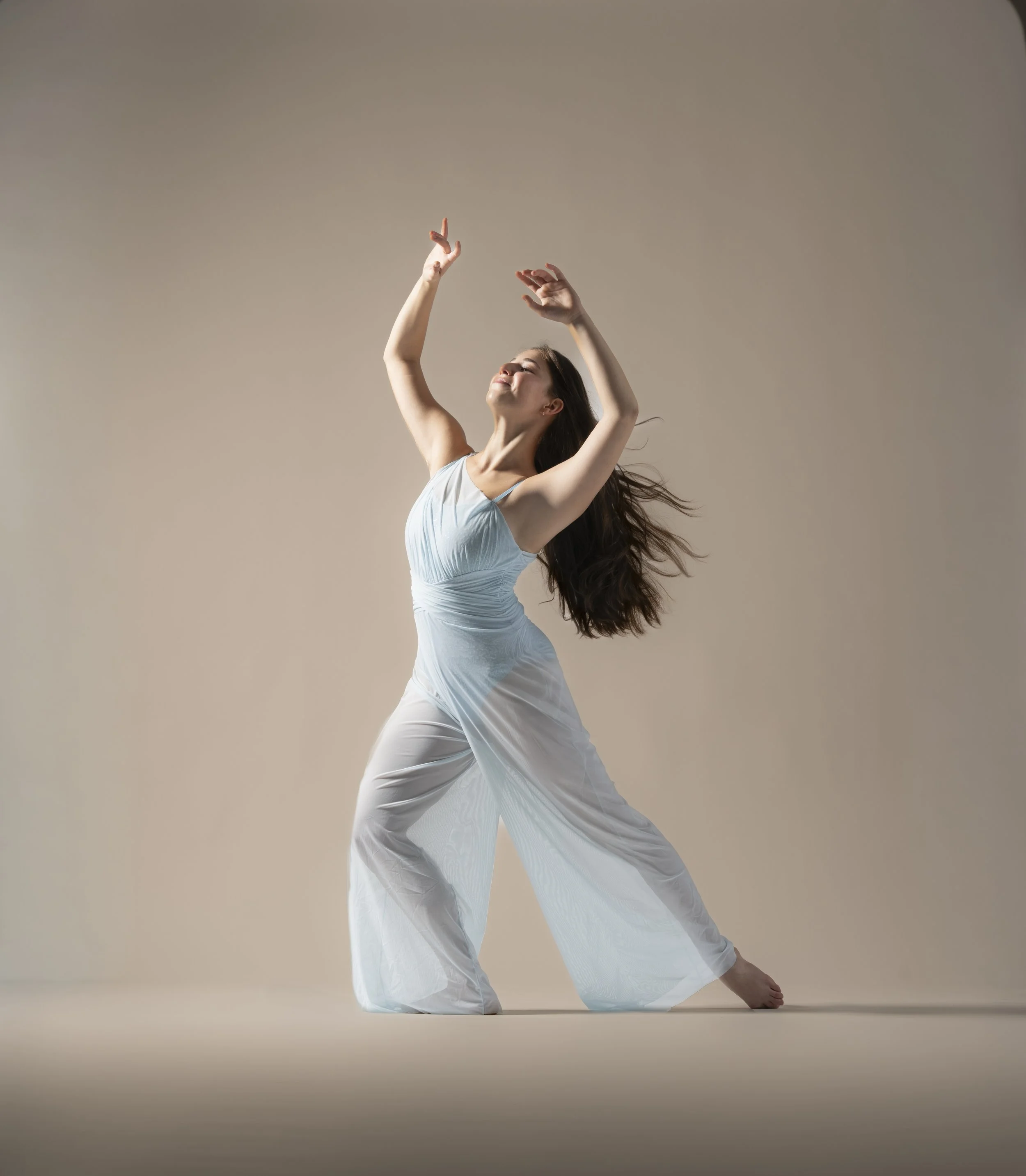 A woman with long brown hair dancing barefoot, wearing a light blue sleeveless top and flowing white pants, performing a contemporary dance pose against a neutral background.