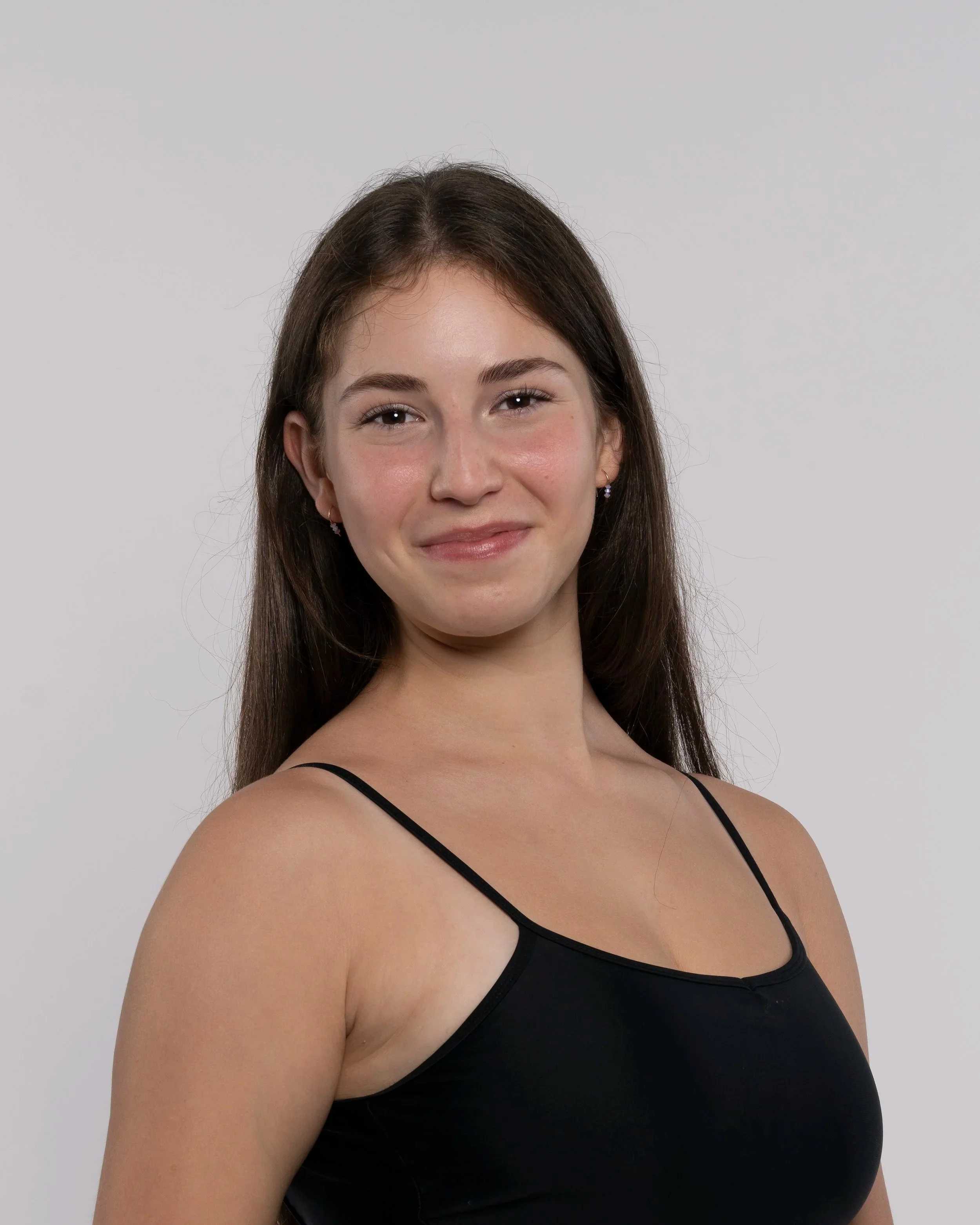 Close-up of a woman with long brown hair wearing a black spaghetti strap top, smiling slightly, against a plain light grey background.