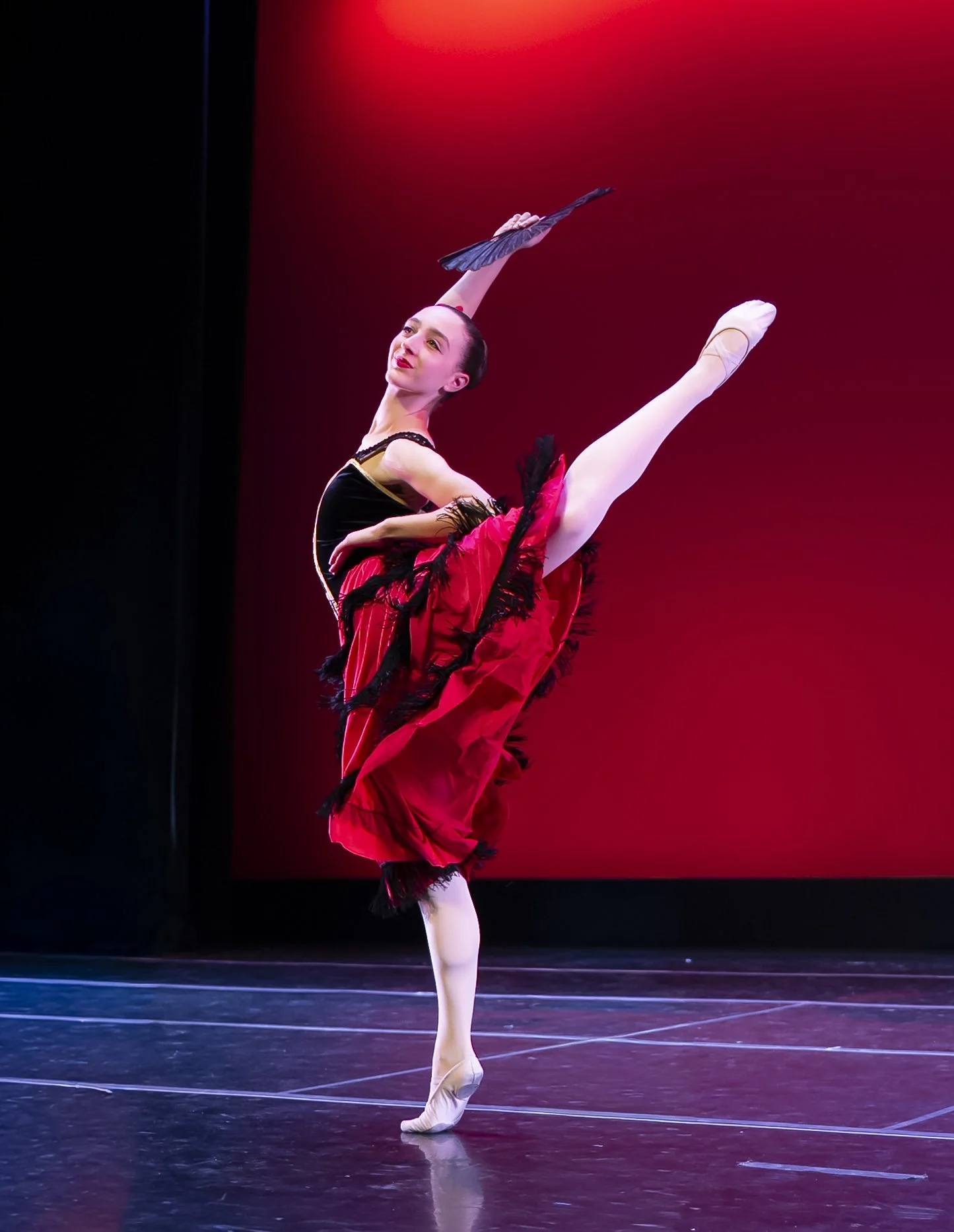 A ballet dancer performs on stage, balancing on one foot with the other leg extended high above her head, holding a black fan, with a red background behind her.