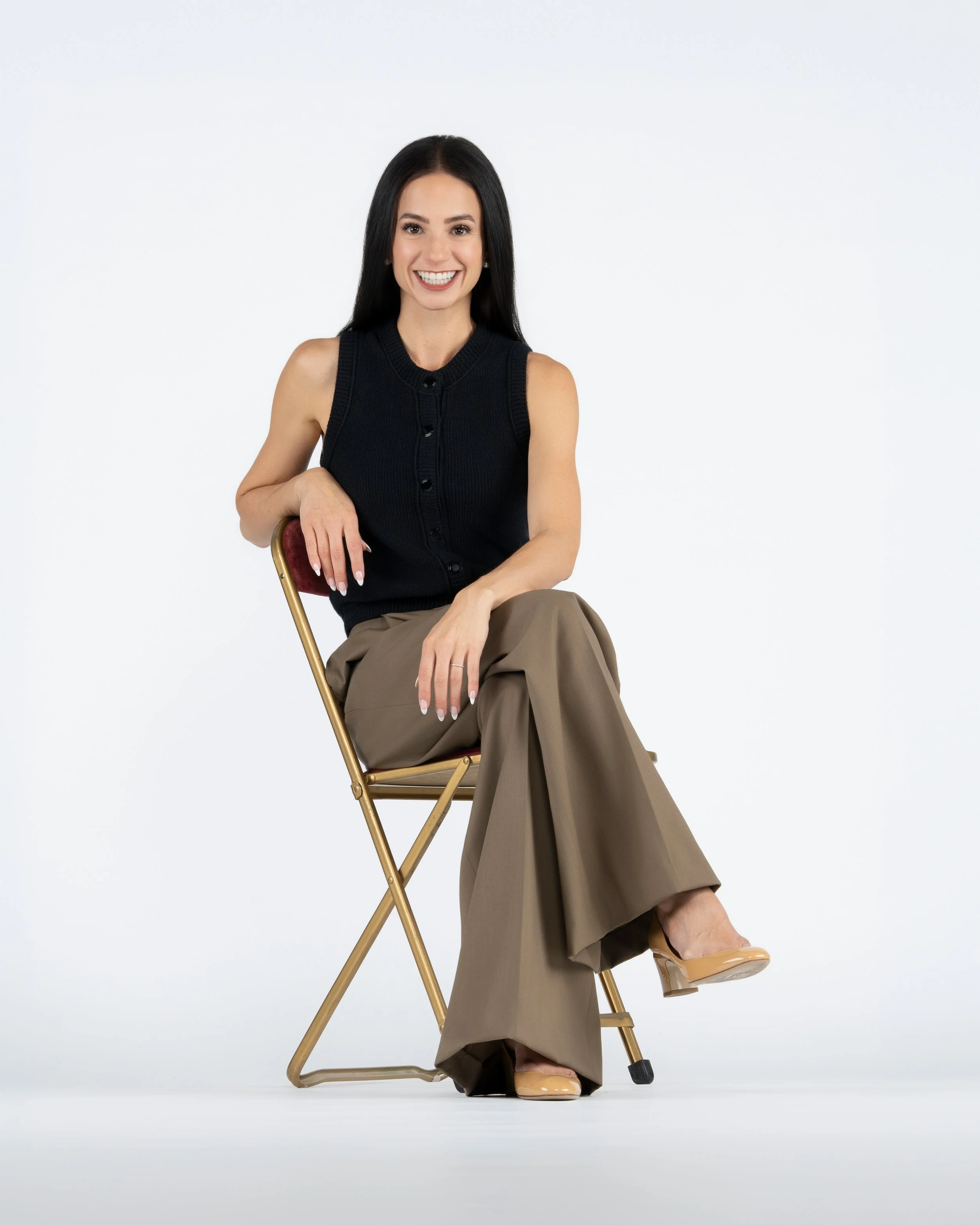 A woman with long dark hair smiling and wearing a black sleeveless top, tan wide-leg pants, and beige shoes, sitting on a gold folding chair against a plain white background.