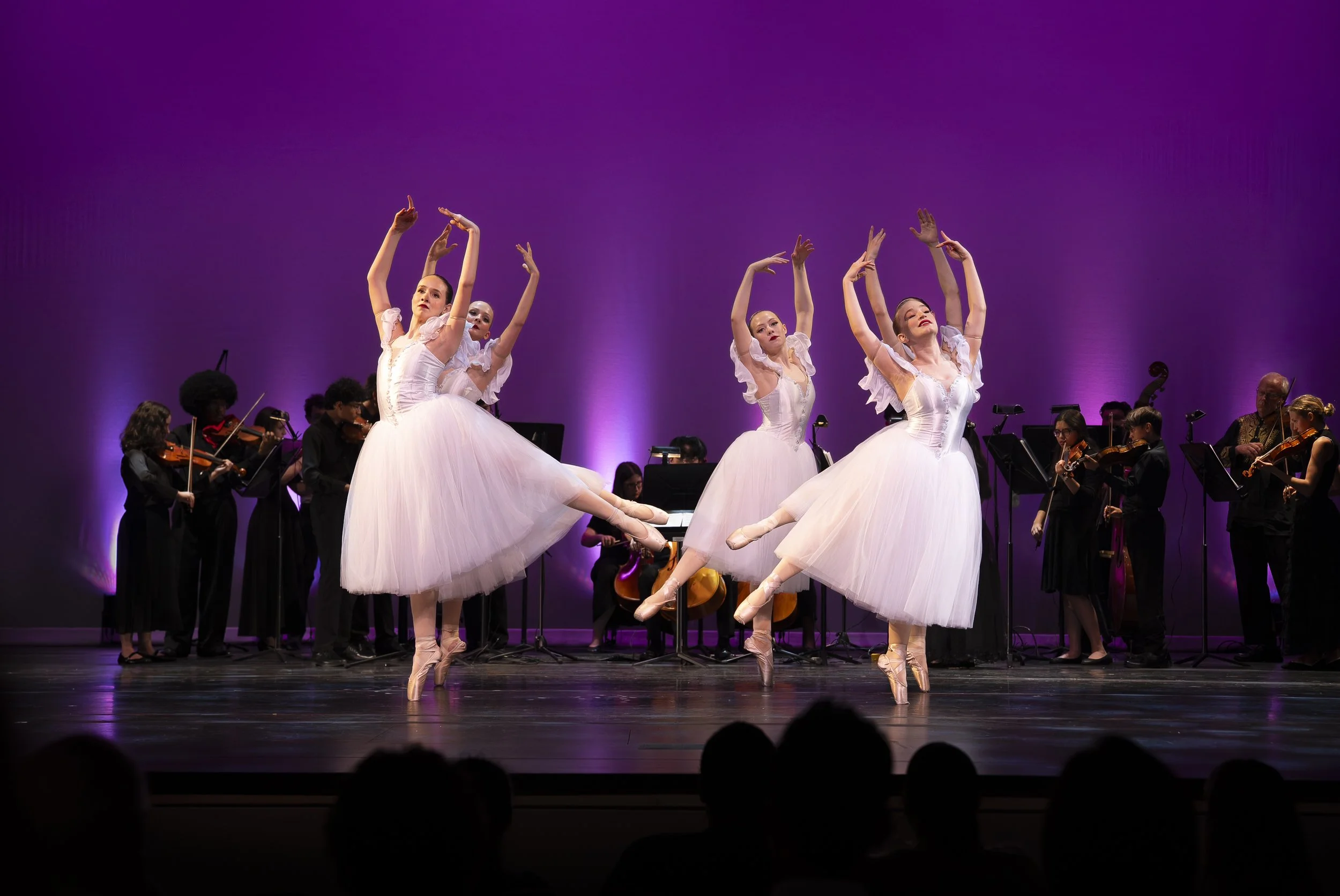 Ballet dancers performing on stage with musicians playing string instruments in the background, purple lighting.