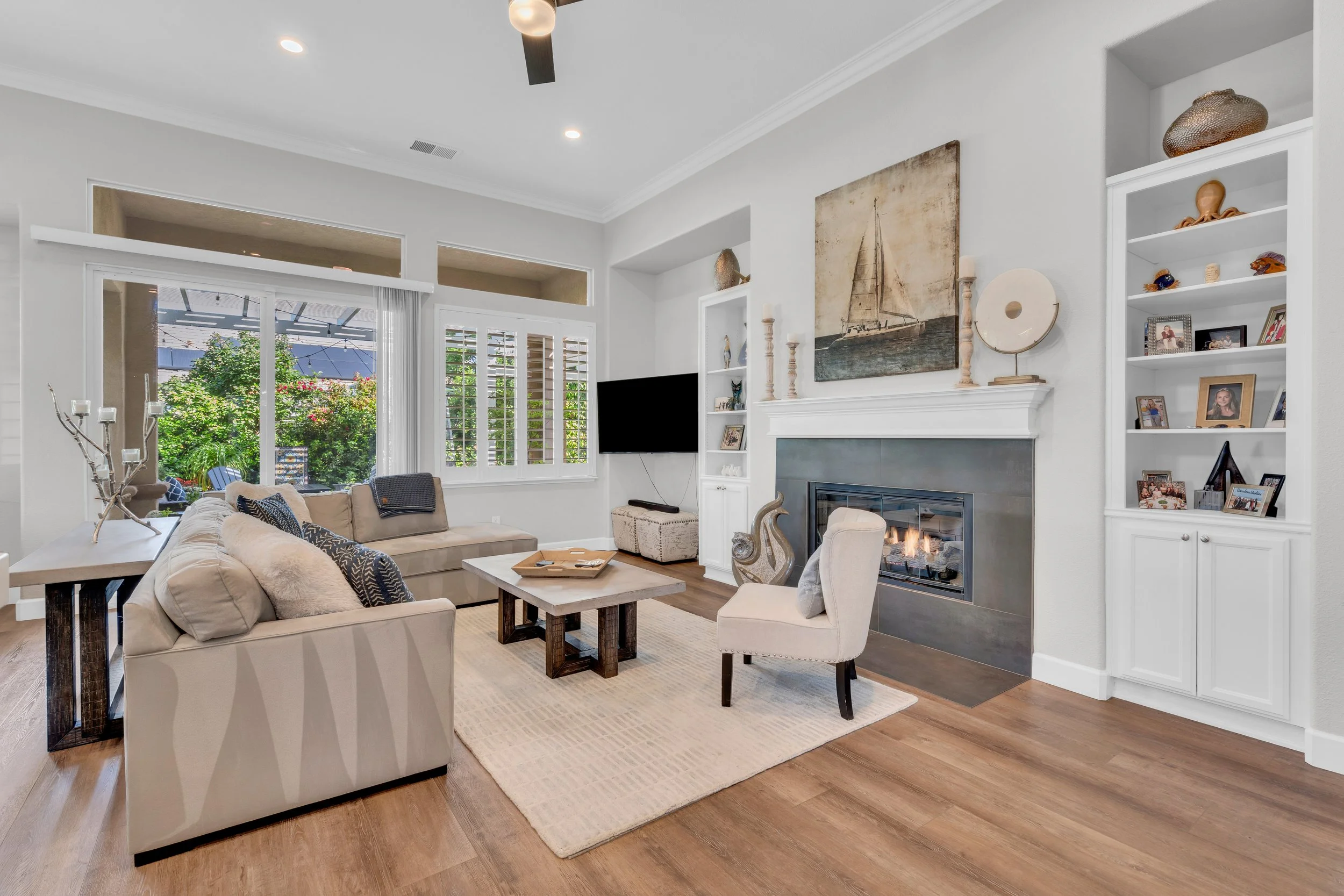 Living room with beige sofa, armchair, coffee table, fireplace, built-in white shelves, and large window with shutters showing outdoor greenery.