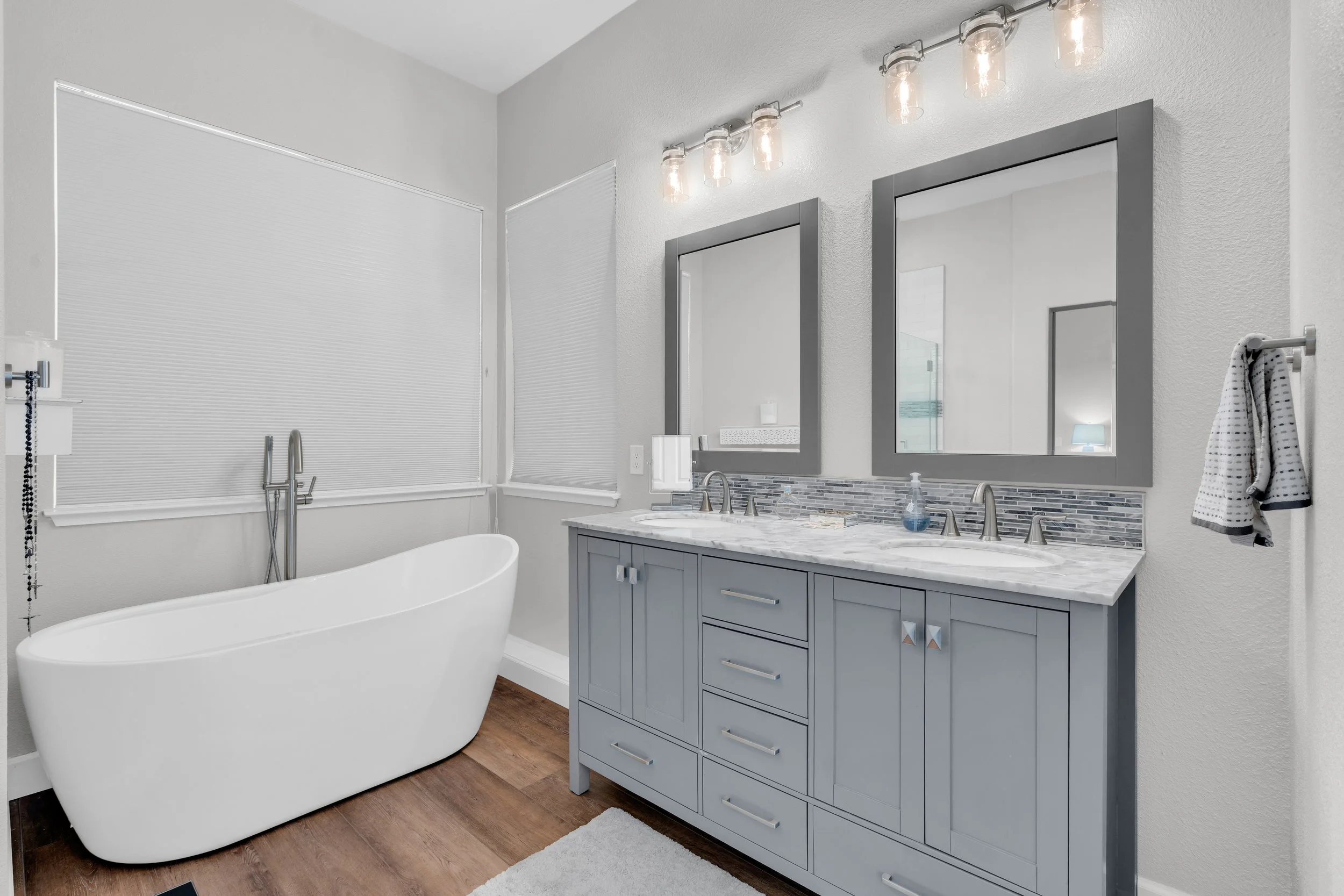 Modern bathroom with a freestanding bathtub, gray vanity with marble countertop, two mirrors with gray frames, and wooden flooring.