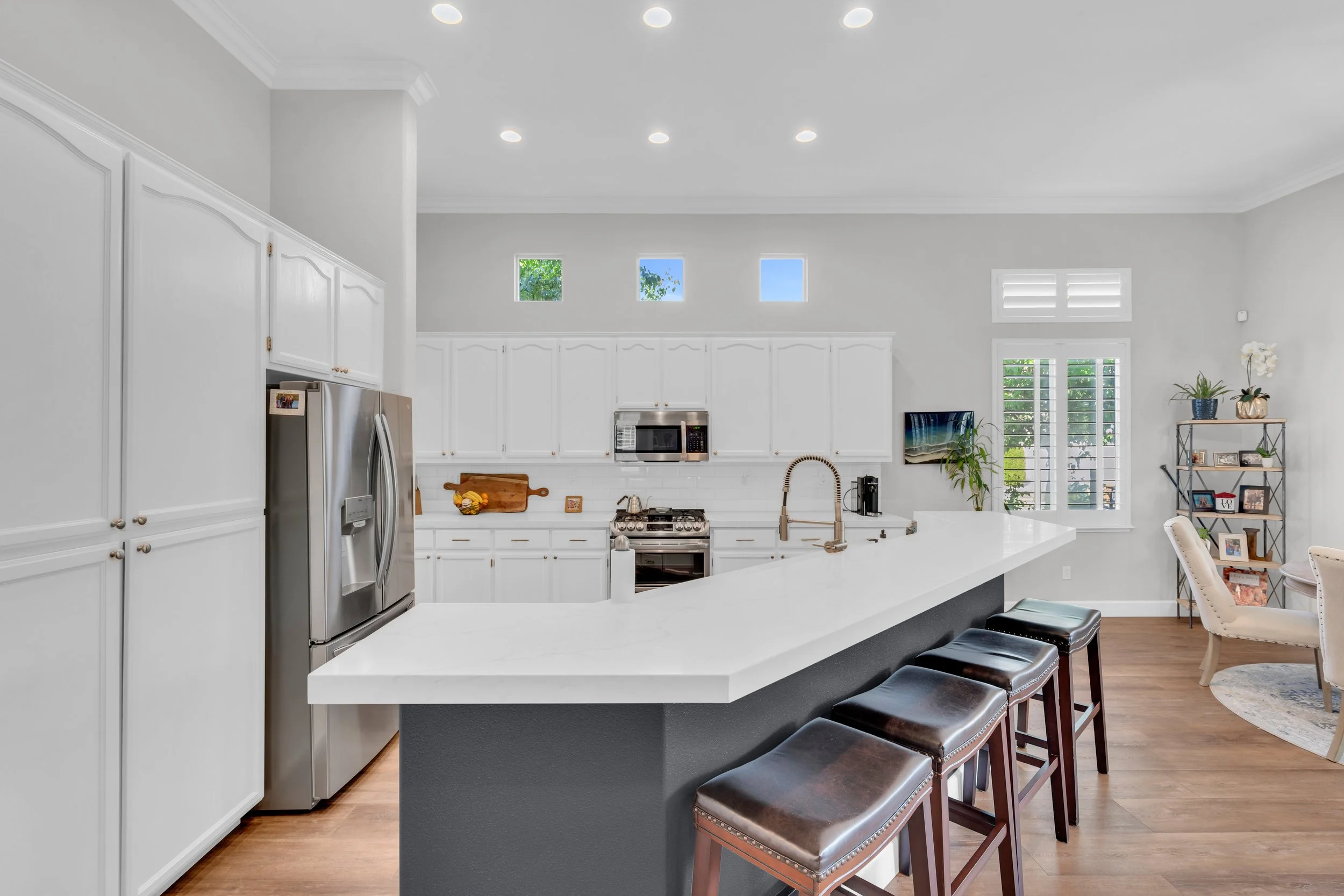 Kitchen with white cabinets, stainless steel refrigerator, microwave, gas stove, and island with bar stools. Bright windows and a small shelf with decorative plants and framed photos.