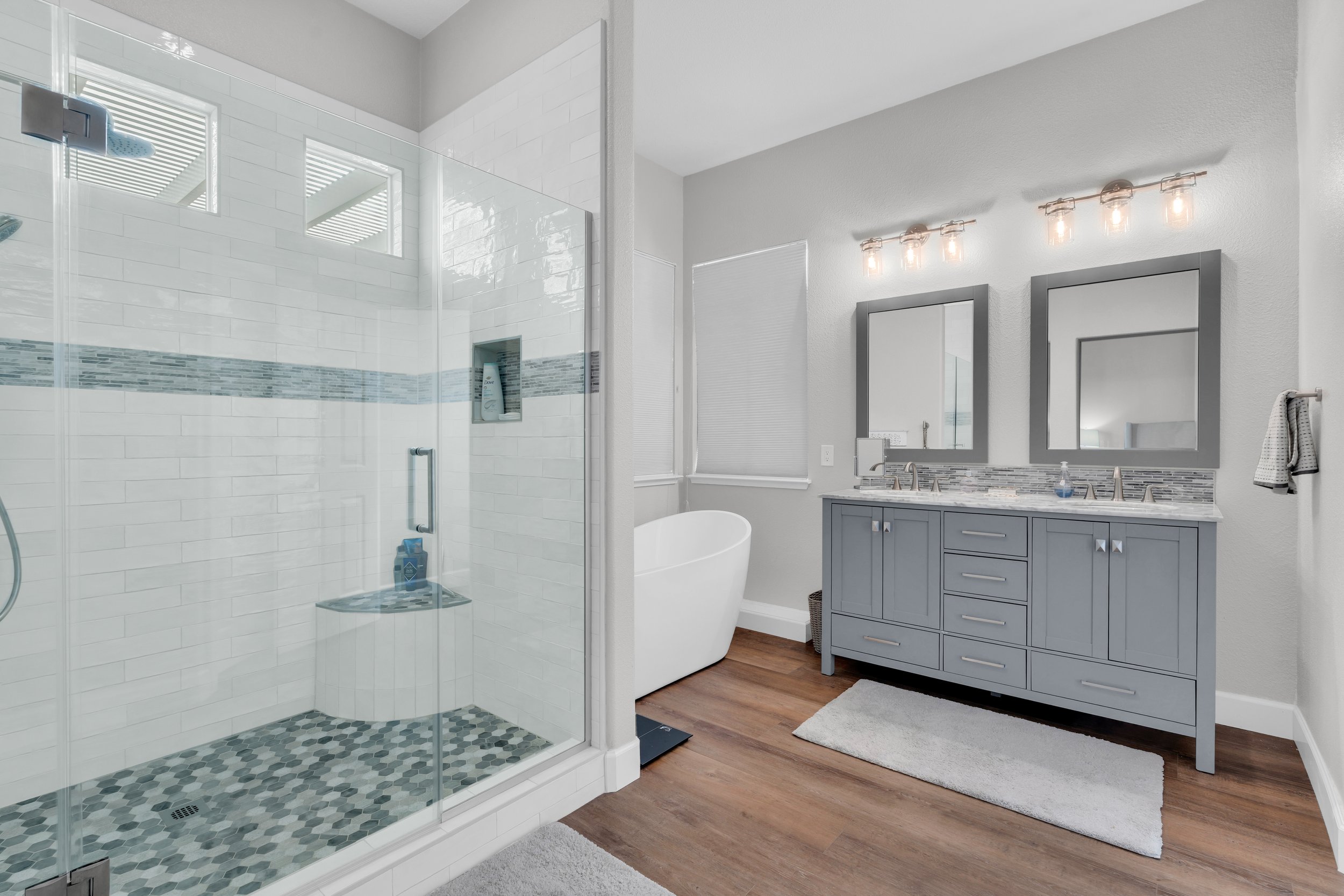 A modern bathroom with a glass-enclosed shower on the left with white tiles and a mosaic tile floor. On the right, there is a gray vanity with a marble countertop, two mirrors, two light fixtures, and a white oval bathtub. The floor is wooden, and there are two windows with blinds.