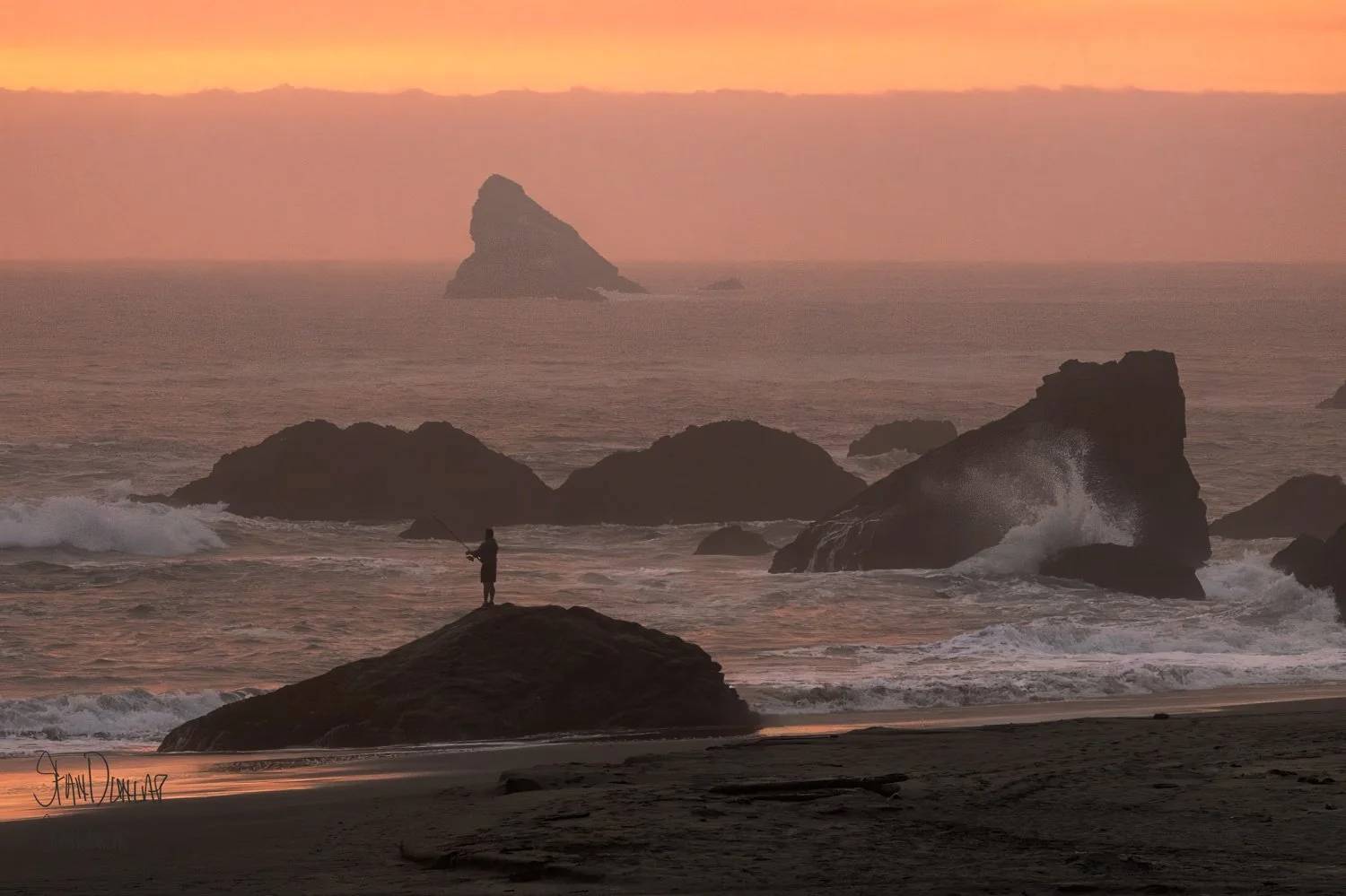 a man surf fishing stands on a large shoreline rock as the surf sprays against rocks during an orange sky sunset