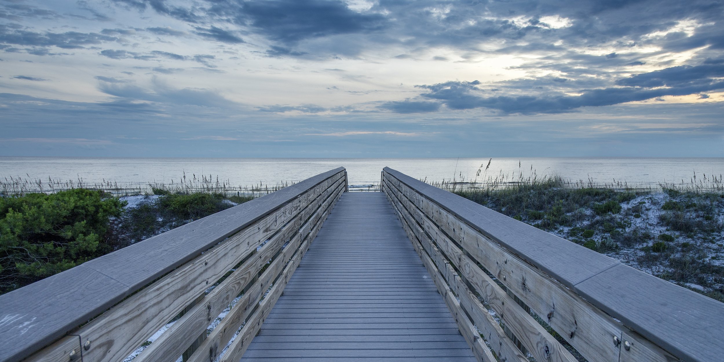 Wooden boardwalk leading through coastal dunes toward a calm beach and ocean beneath dramatic clouds at sunrise.