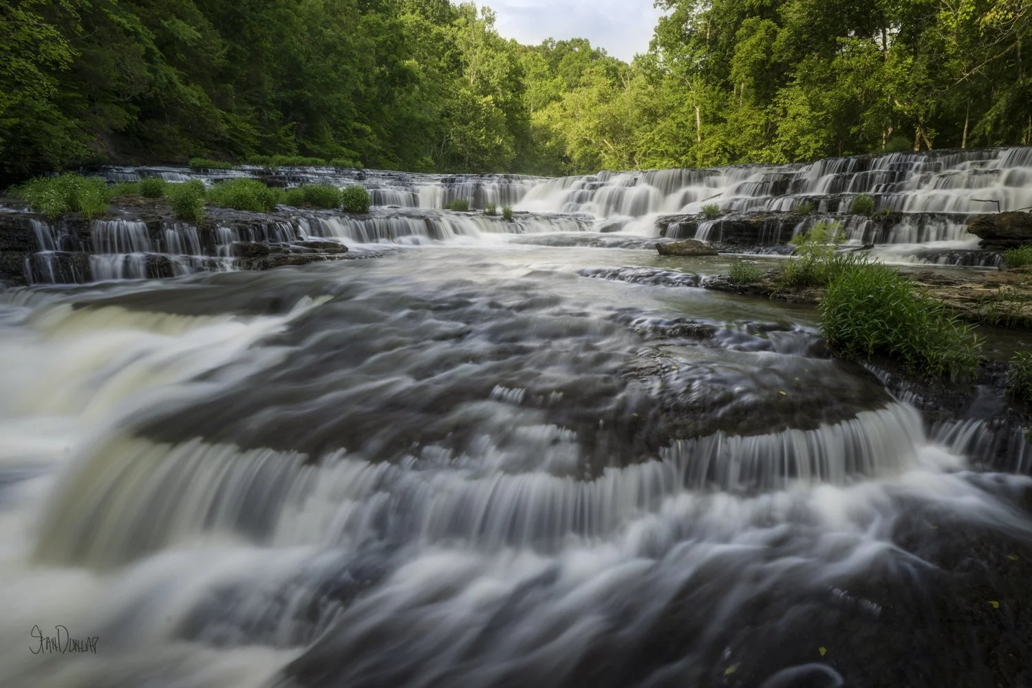 Water cascades over layered rock ledges at Burgess Falls surrounded by summer greenery.