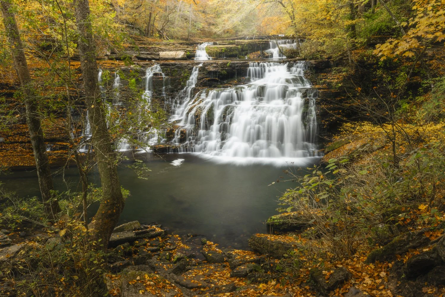 The white waters of Rutledge Falls flow into a green pool surrounded by colorful fall leaves.