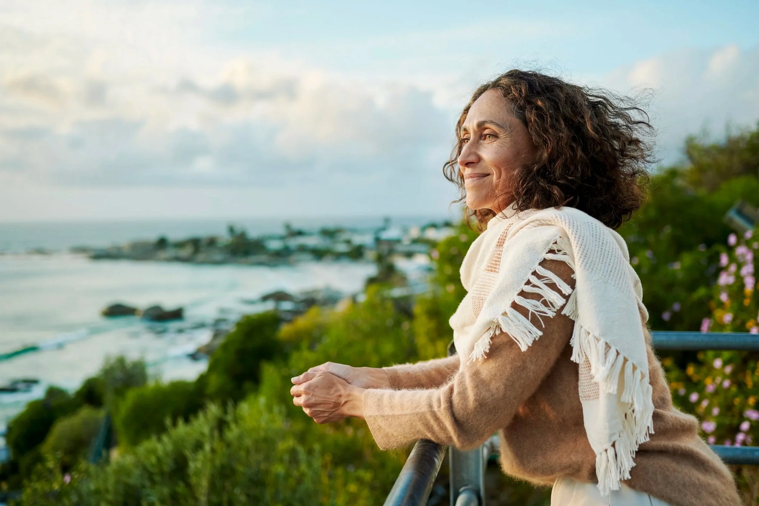 A woman with curly hair smiling and looking into the distance while leaning on a railing outdoors near the coast, with greenery and a cloudy sky in the background.