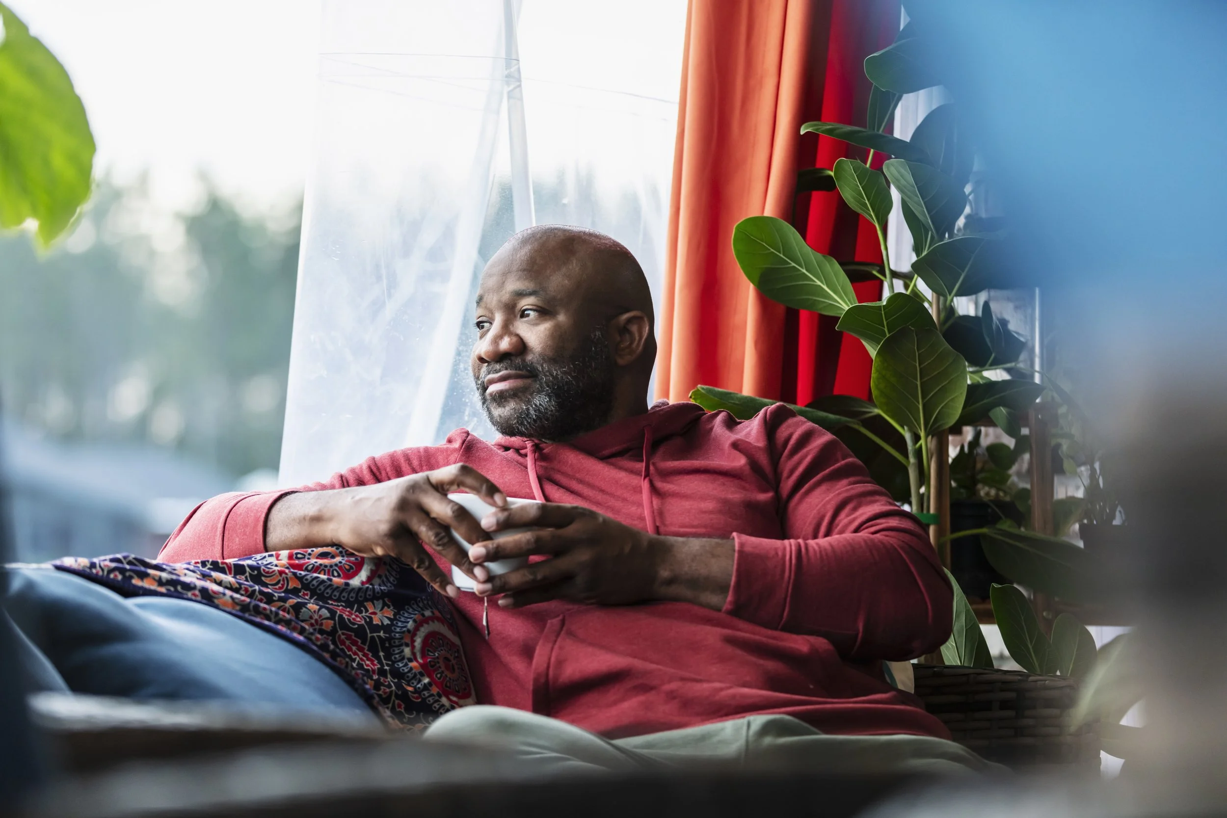 A bald man with a beard wearing a red hoodie, sitting indoors near a window with orange curtains and green plants, holding a smartphone and looking out thoughtfully.