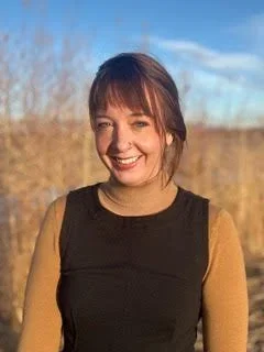 A smiling woman with brown hair, wearing a black sleeveless top over a tan long-sleeve shirt, standing outdoors with a blue sky and trees in the background.
