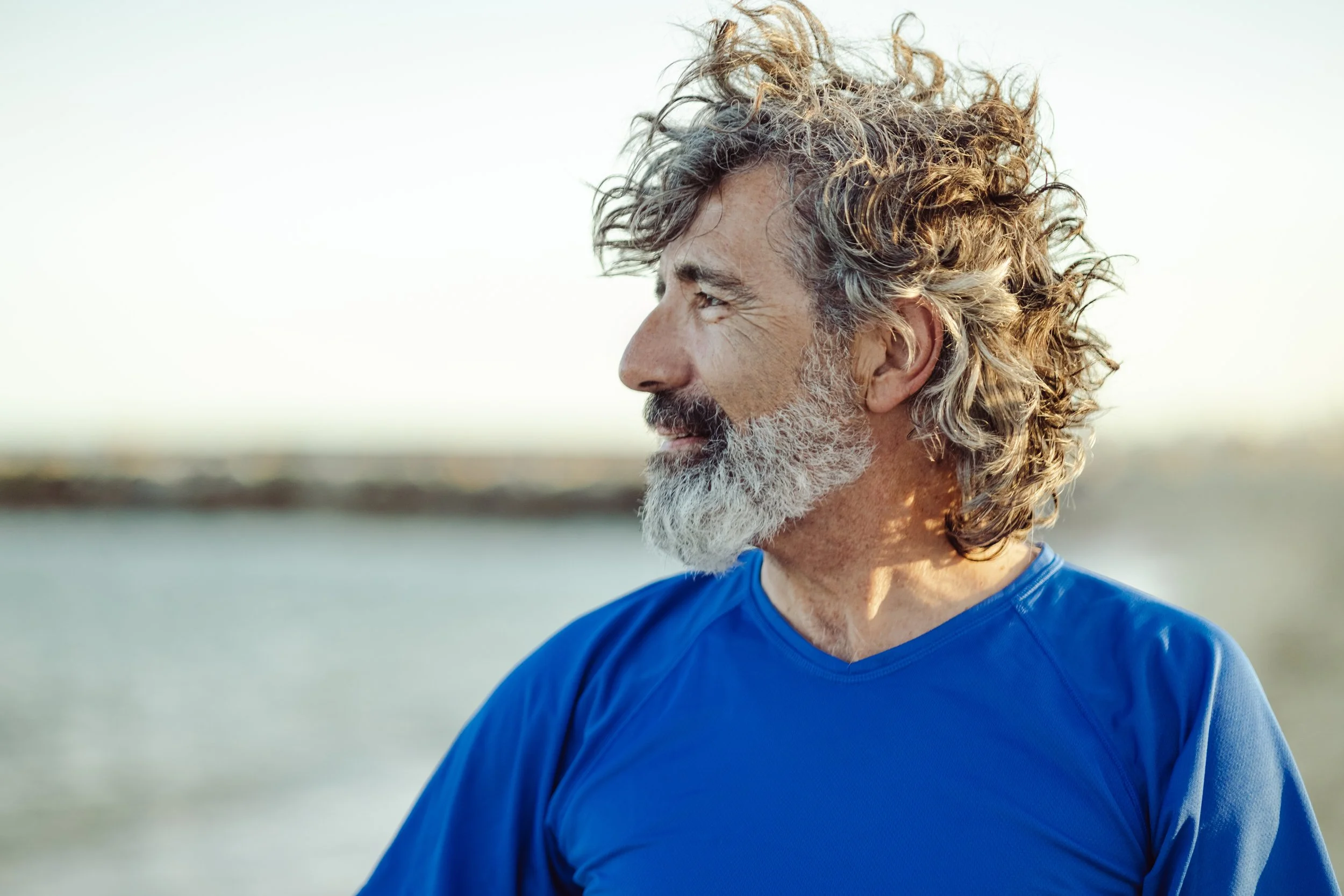 Side profile of an elderly man with curly gray hair and a beard, wearing a blue shirt, outdoors near water.