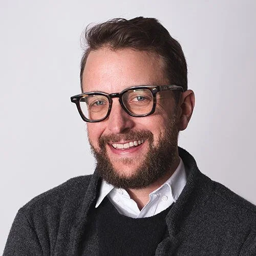 A smiling man with glasses, facial hair, and short brown hair wearing a white shirt and dark sweater against a plain light background.