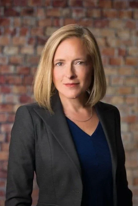 A woman with shoulder-length blonde hair wearing a black blazer and blue top, standing in front of a brick wall.
