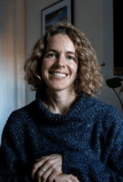 A woman with curly hair smiling indoors, wearing a dark blue sweater, with a picture frame and a plant in the background.