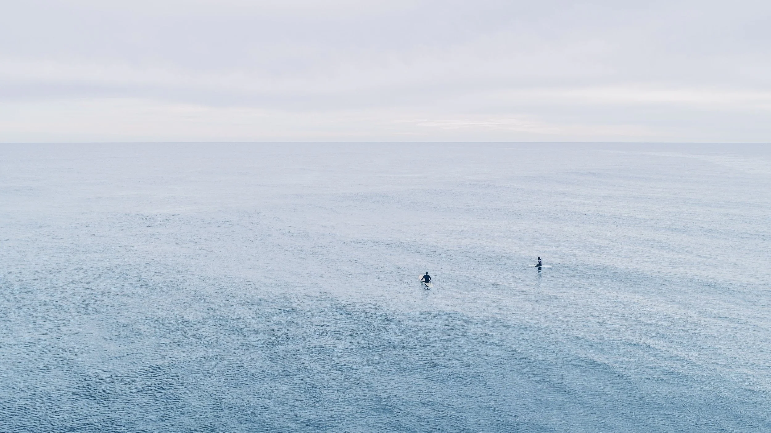 Two surfers standing on surfboards in the ocean, waiting for waves under a cloudy sky.