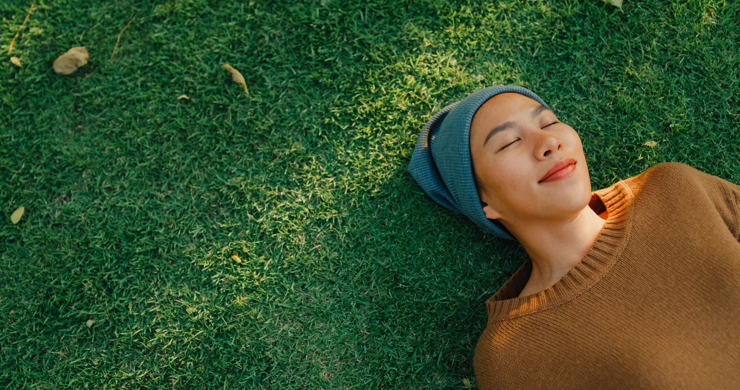 Young woman lying on grass with eyes closed, wearing a brown sweater and a gray beanie, smiling softly.