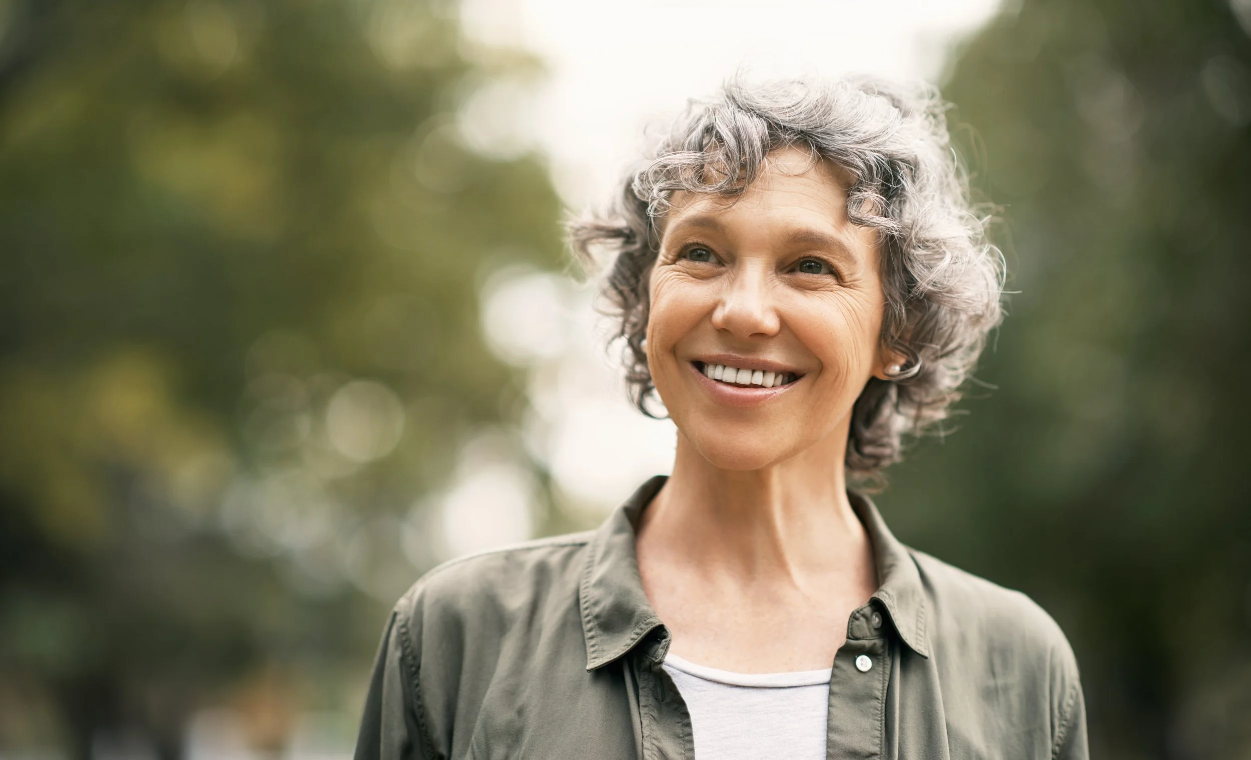A smiling woman with short, curly gray hair standing outdoors with greenery and sunlight in the background.