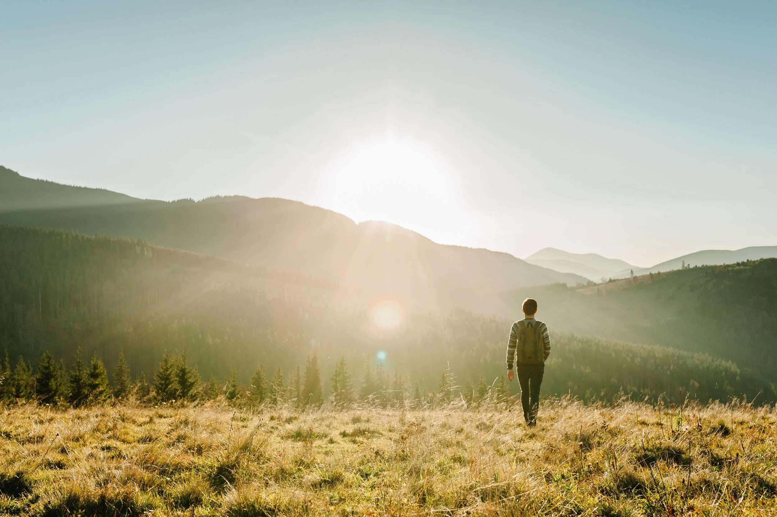 A person wearing a backpack is standing on a grassy field, facing mountains with the sun shining brightly behind them, creating a lens flare.