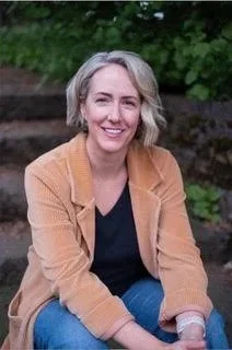 Smiling woman with shoulder-length gray hair, wearing a tan blazer over a black shirt, sitting outdoors with greenery in the background.