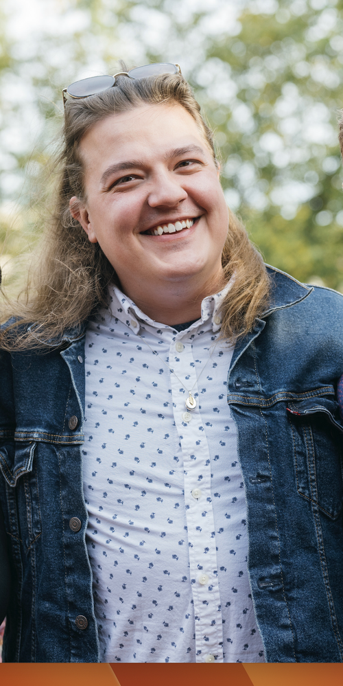 A smiling person with long light brown hair wearing sunglasses on their head, a patterned button-up shirt, and a denim jacket outdoors.
