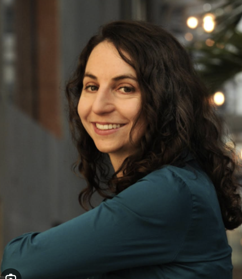 A woman with dark curly hair smiling and looking at the camera. She is wearing a teal top and is indoors near a window with blurred lights in the background.