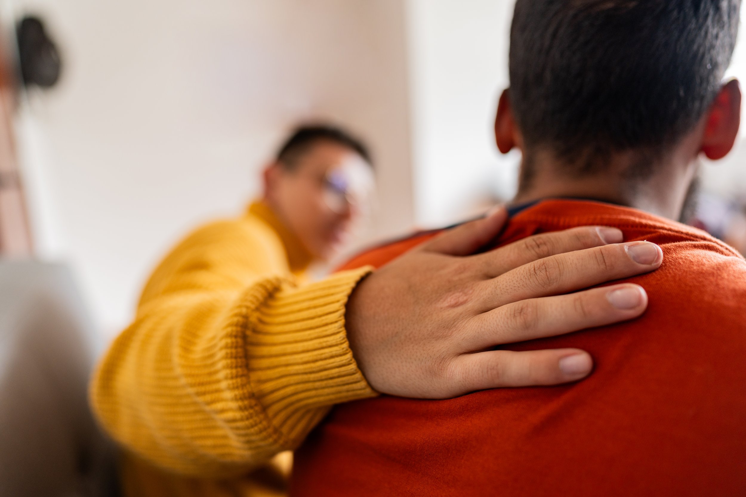 Close-up of two people hugging, one with a hand on the other's shoulder, wearing a yellow sweater and an orange shirt.