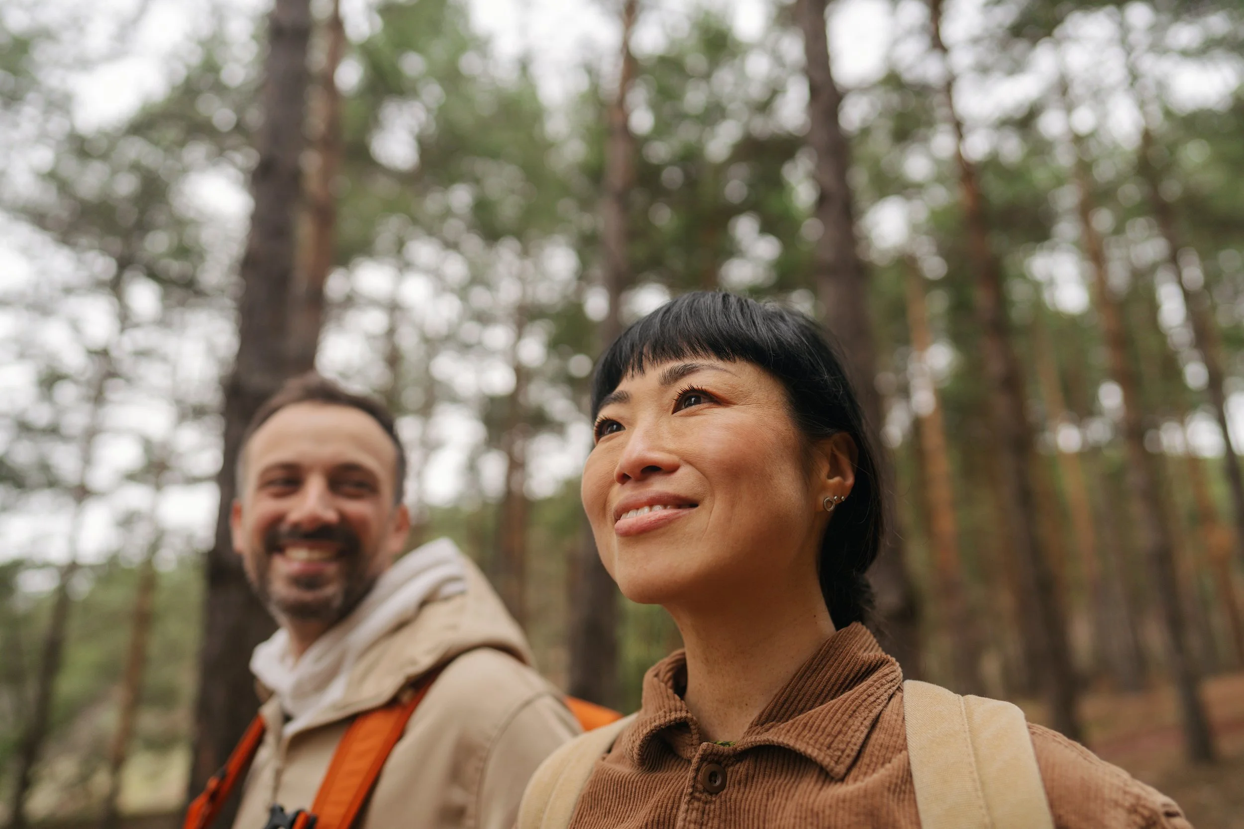 Close-up of two smiling friends with dark hair, one with short hair and the other with slightly longer hair, in a forest with tall trees and green foliage in the background.