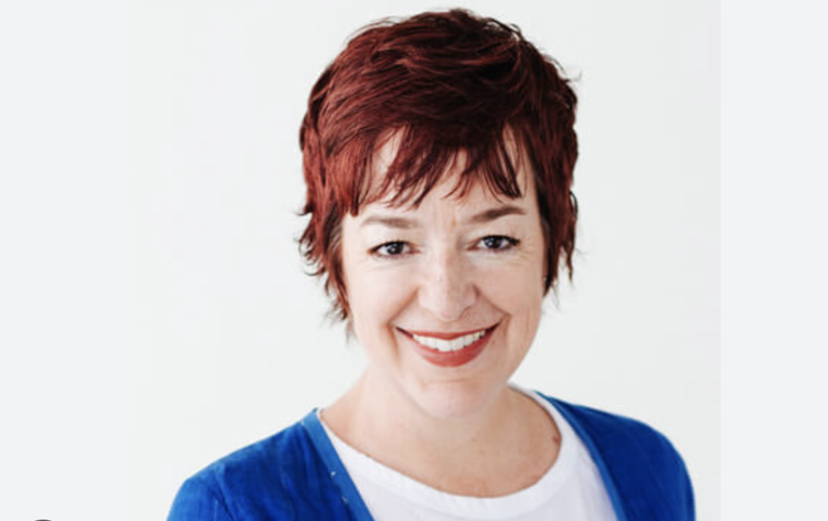 Close-up of a woman with short red hair, smiling, wearing a blue cardigan over a white shirt, against a plain background.
