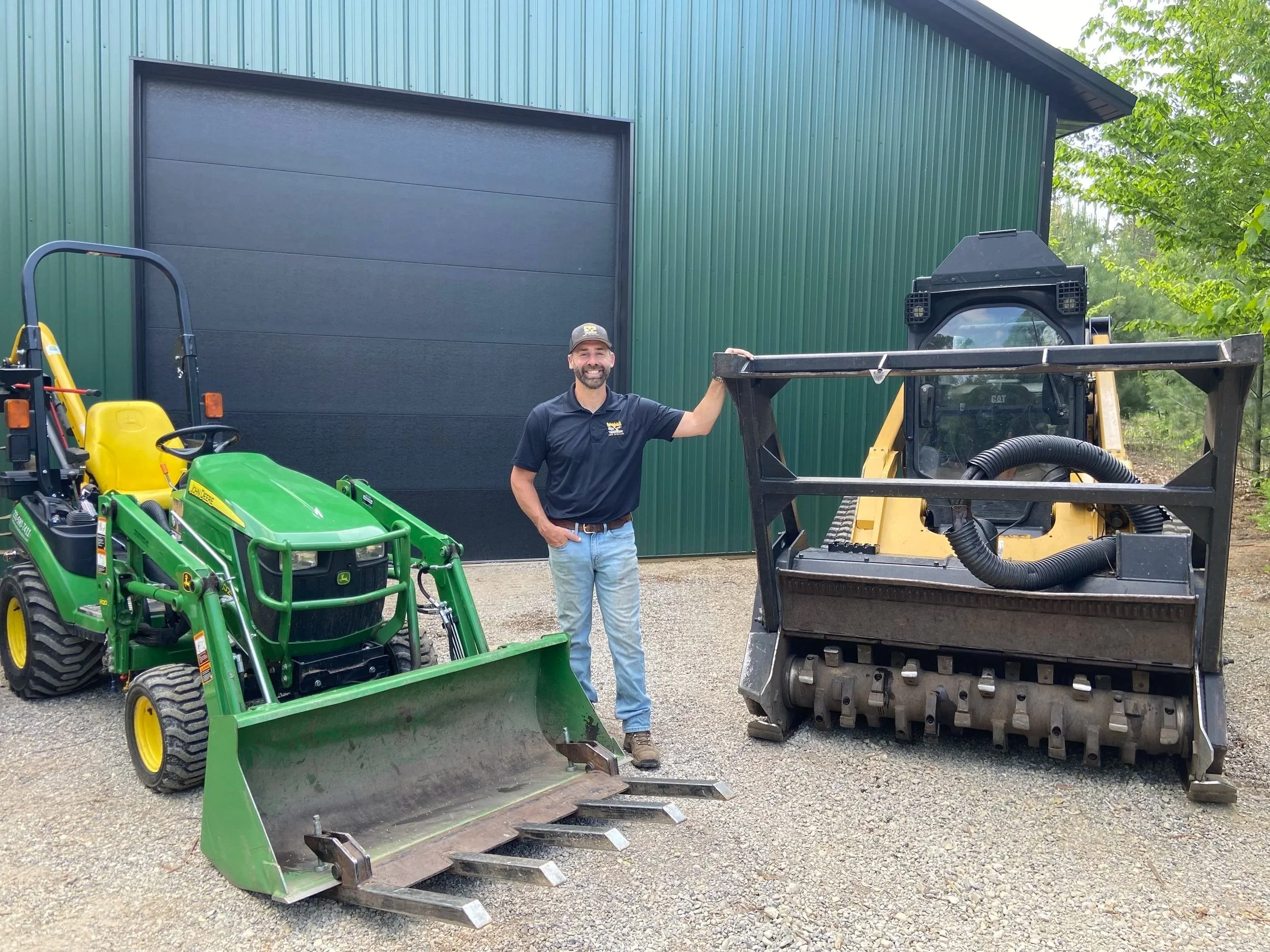 A man smiling and standing next to construction equipment outside a green metal building surrounded by trees.