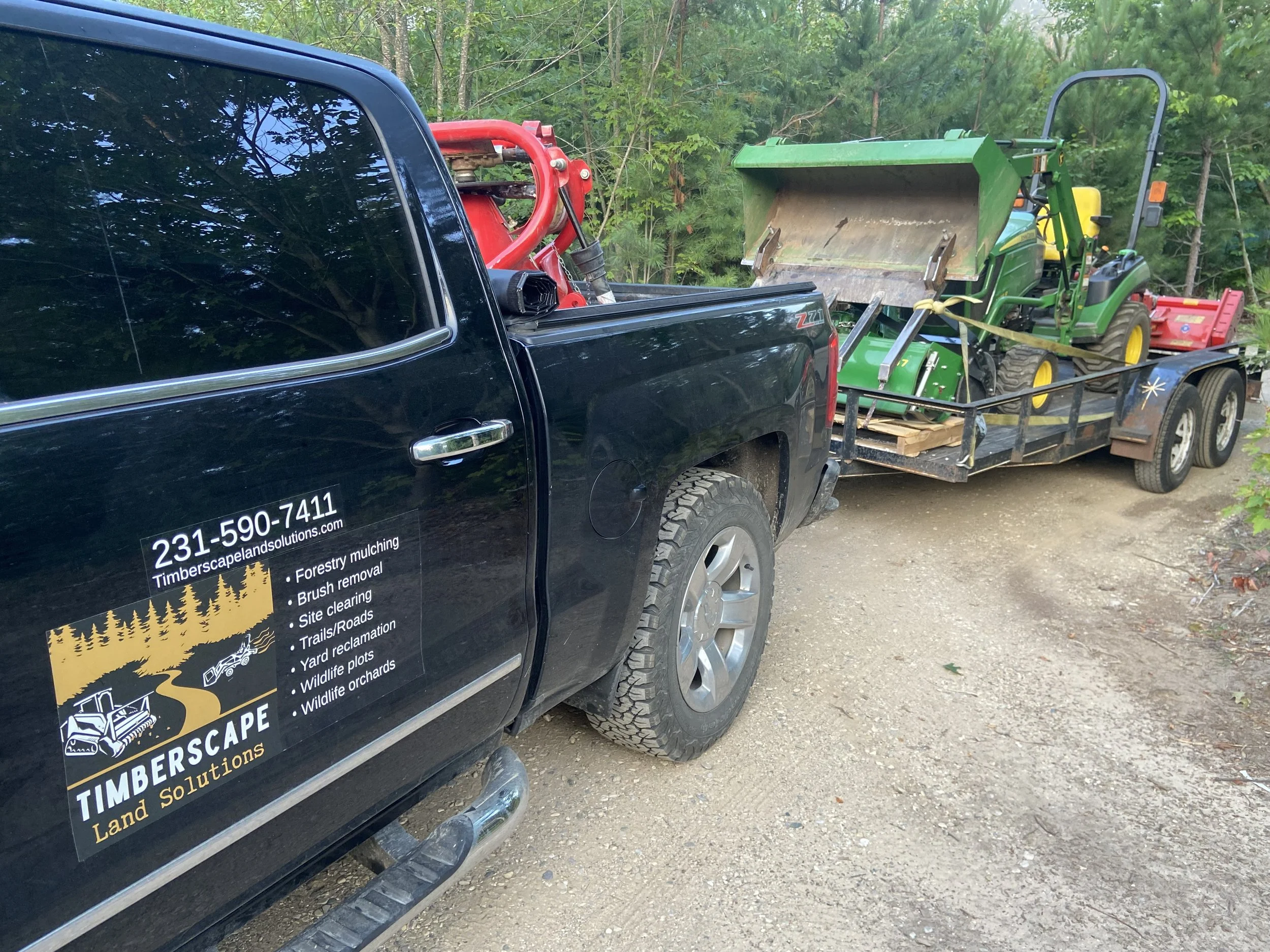 A black pickup truck parked on a dirt road with a trailer attached. The trailer is loaded with a green tractor and a red attachment. The side of the truck displays the text '231-590-7411 TimberScap Land Solutions' with a list of landscaping and forestry services.